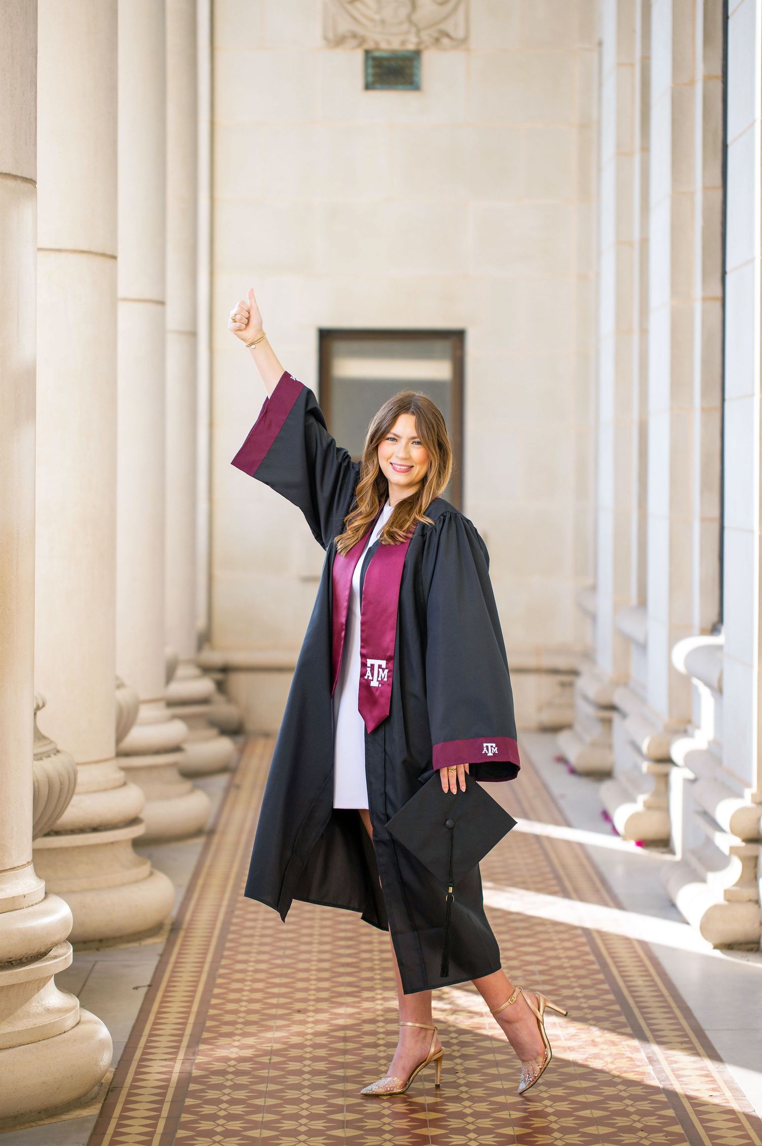 A woman in a black graduation gown stands in a hallway with columns, holding her diploma.