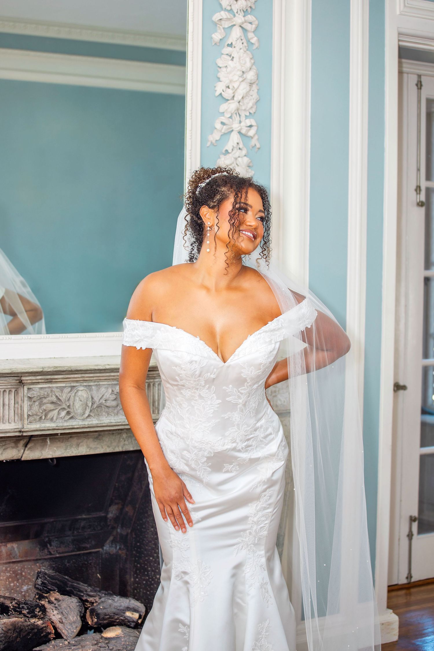Bride in a Coreena's Bridal off-shoulder white lace gown poses in elegant blue-walled room.