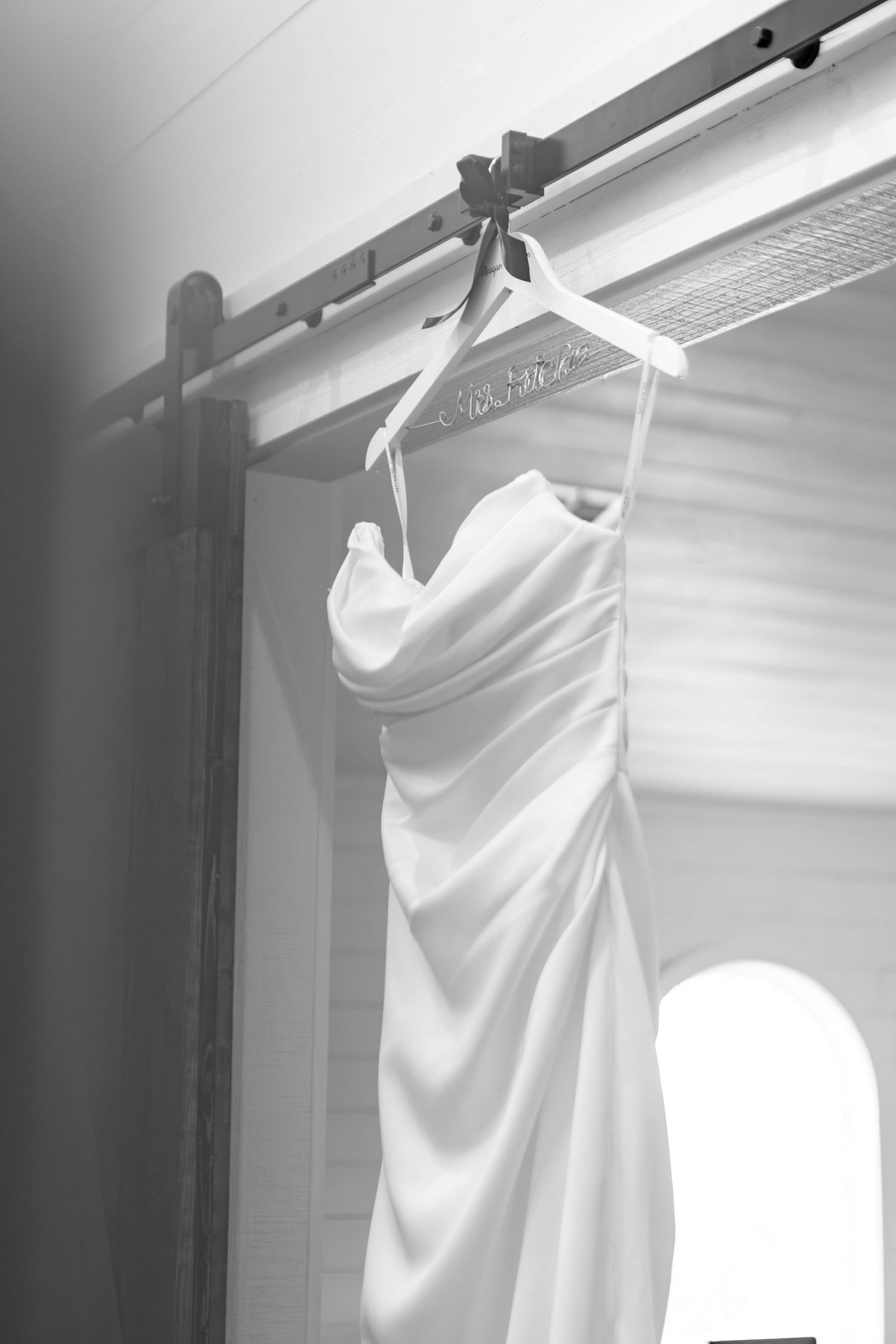 Elegant white wedding dress hanging on a personalized hanger against a window in black and white photography.