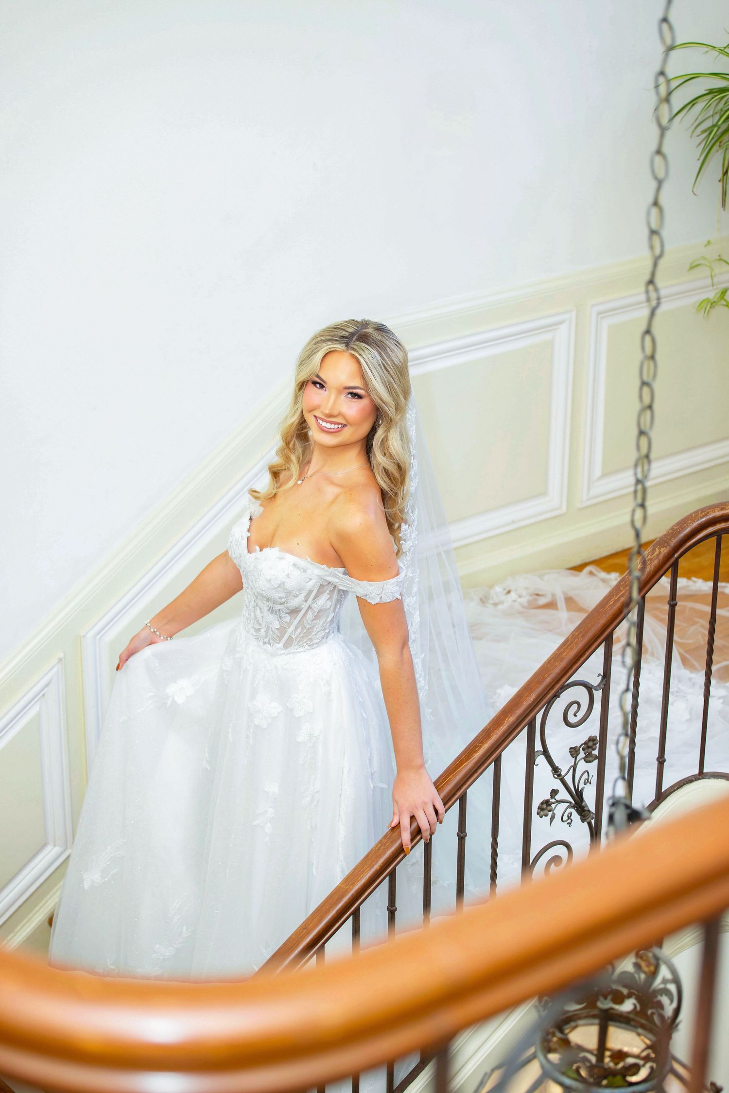 Bride in Coreena's Bridal flows white gown poses gracefully on elegant staircase with wooden railing.