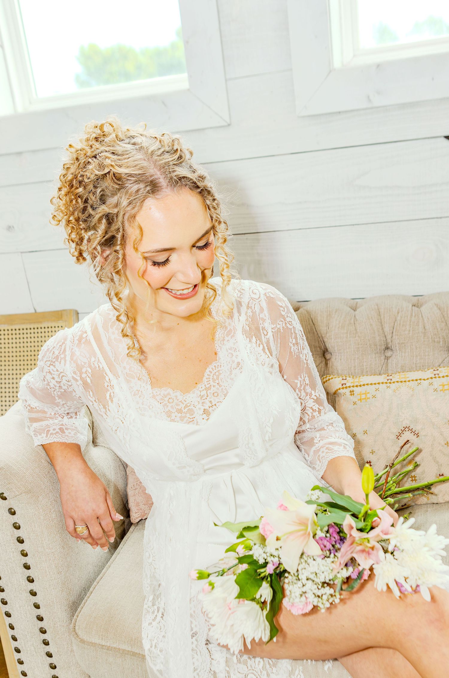 Smiling bride in white lace robe sitting on a couch holding a pink and white floral bouquet.