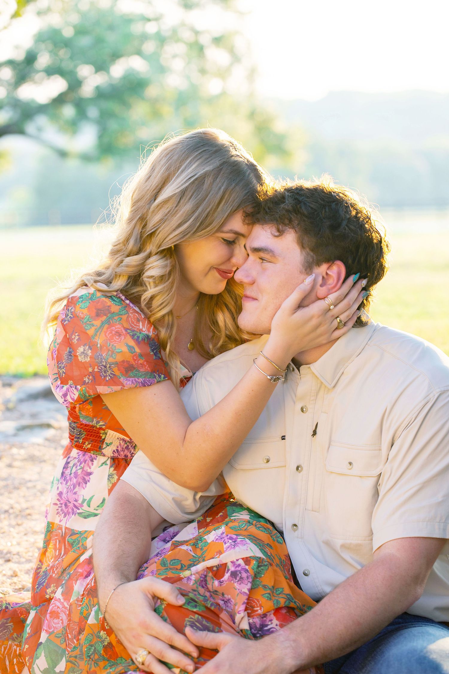 A couple shares intimate moments in a sunlit outdoor setting wearing floral dress and light colored shirt.
