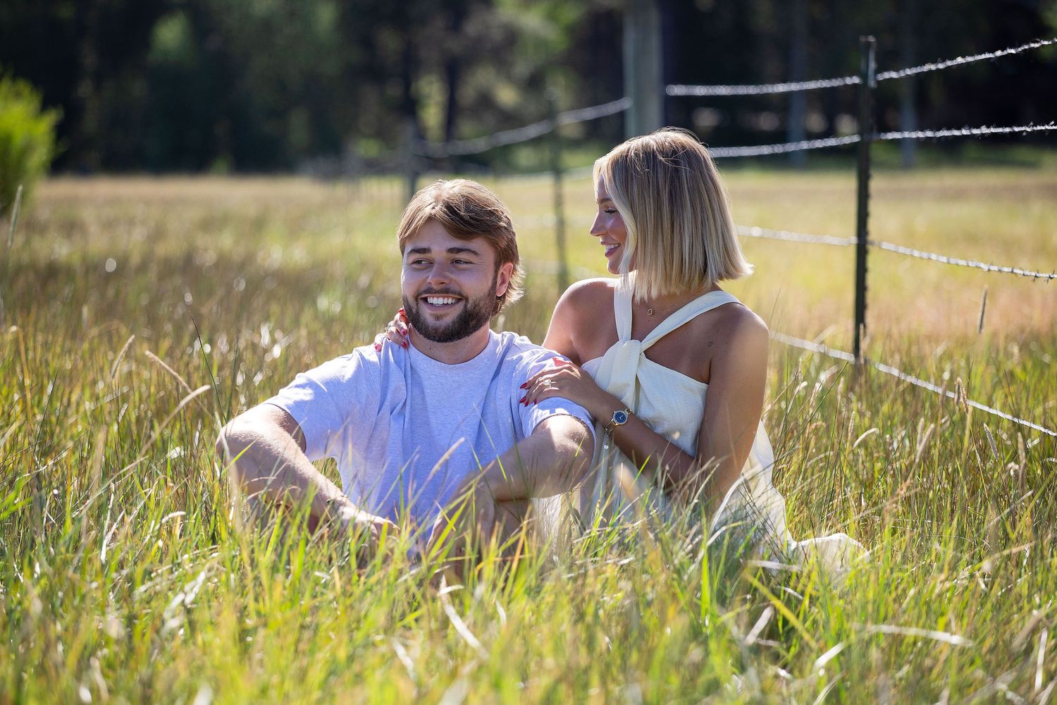 A smiling couple sits together in a sunny grassy field near a barbed wire fence, enjoying a relaxed outdoor moment.