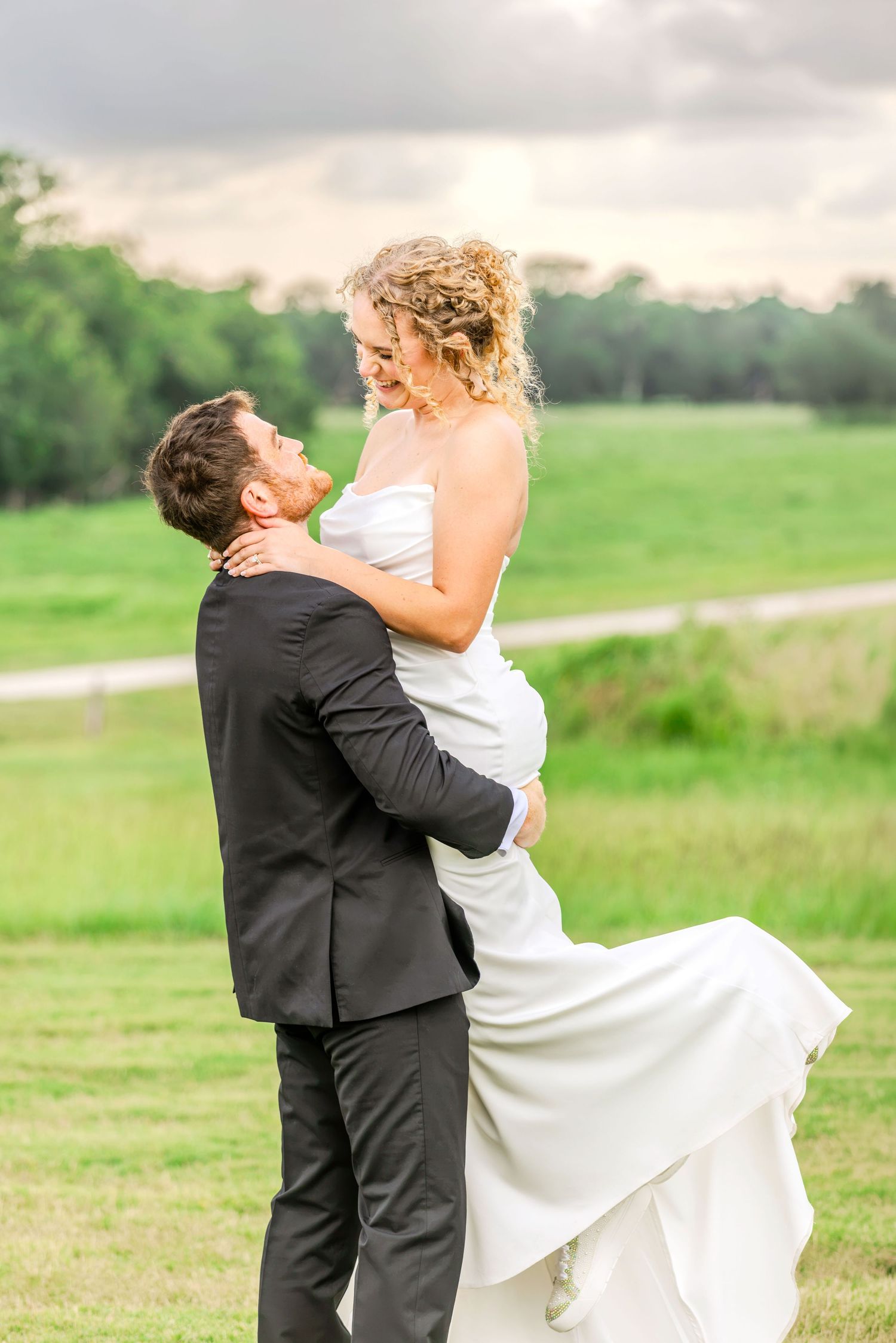 Groom in black suit lifts smiling bride in white wedding dress outdoors on green grass field at sunset.