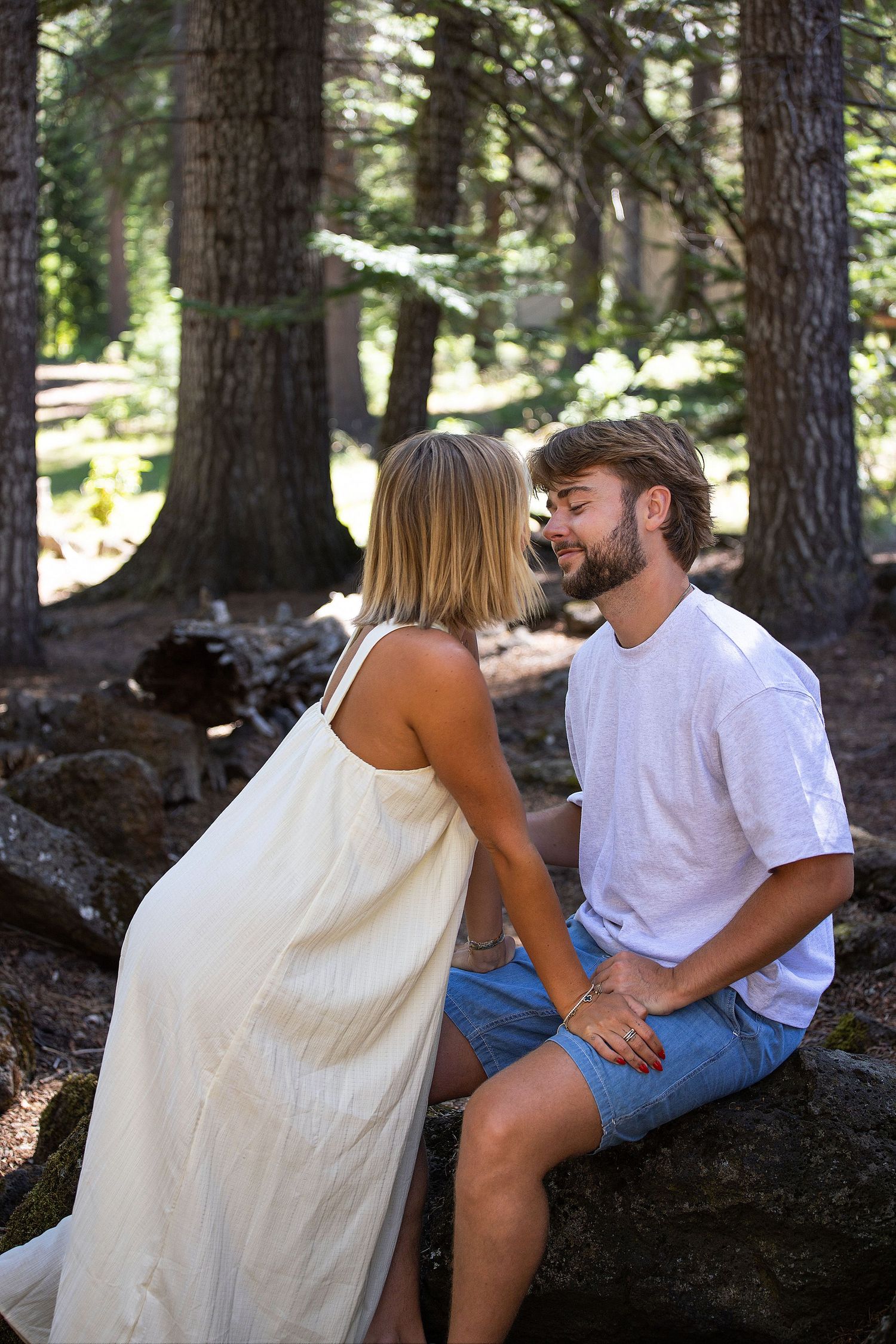A couple shares a romantic moment in a sunlit forest, the woman in a white dress facing a bearded man in a white tee.