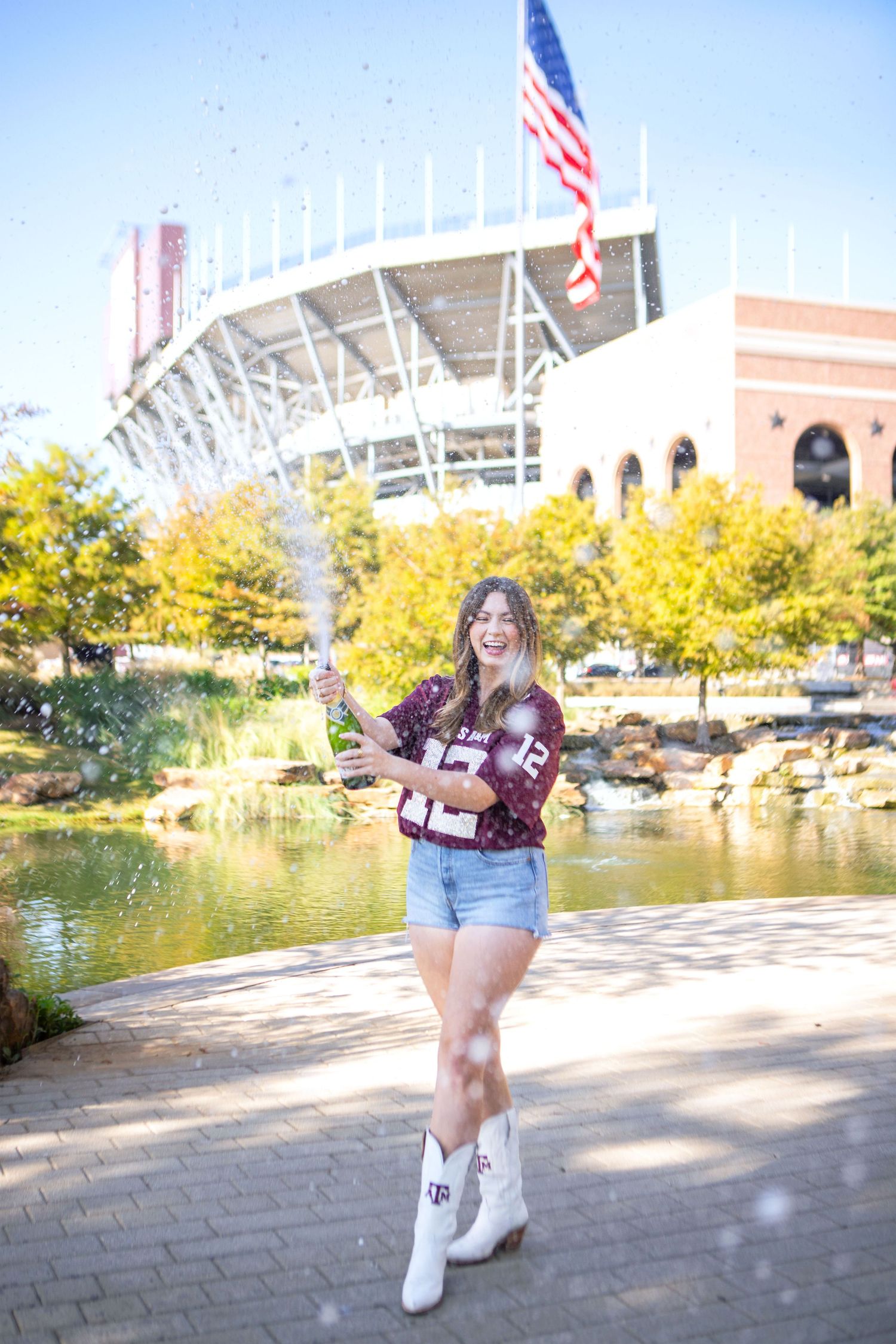 A young woman standing in front of a large sports stadium, holding a football and wearing a maroon jersey.