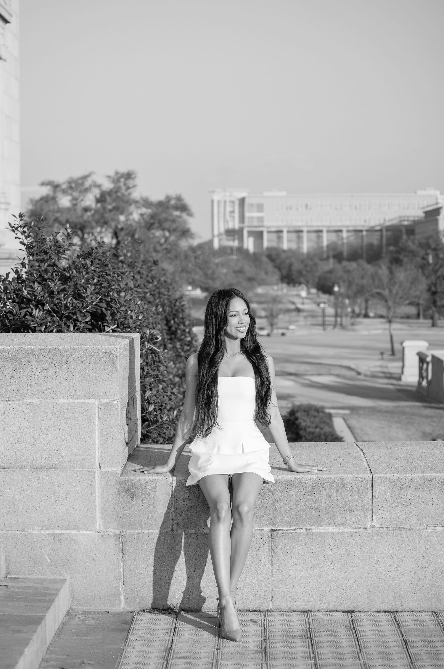 A person sits on a concrete wall outdoors with trees and buildings visible in the background.