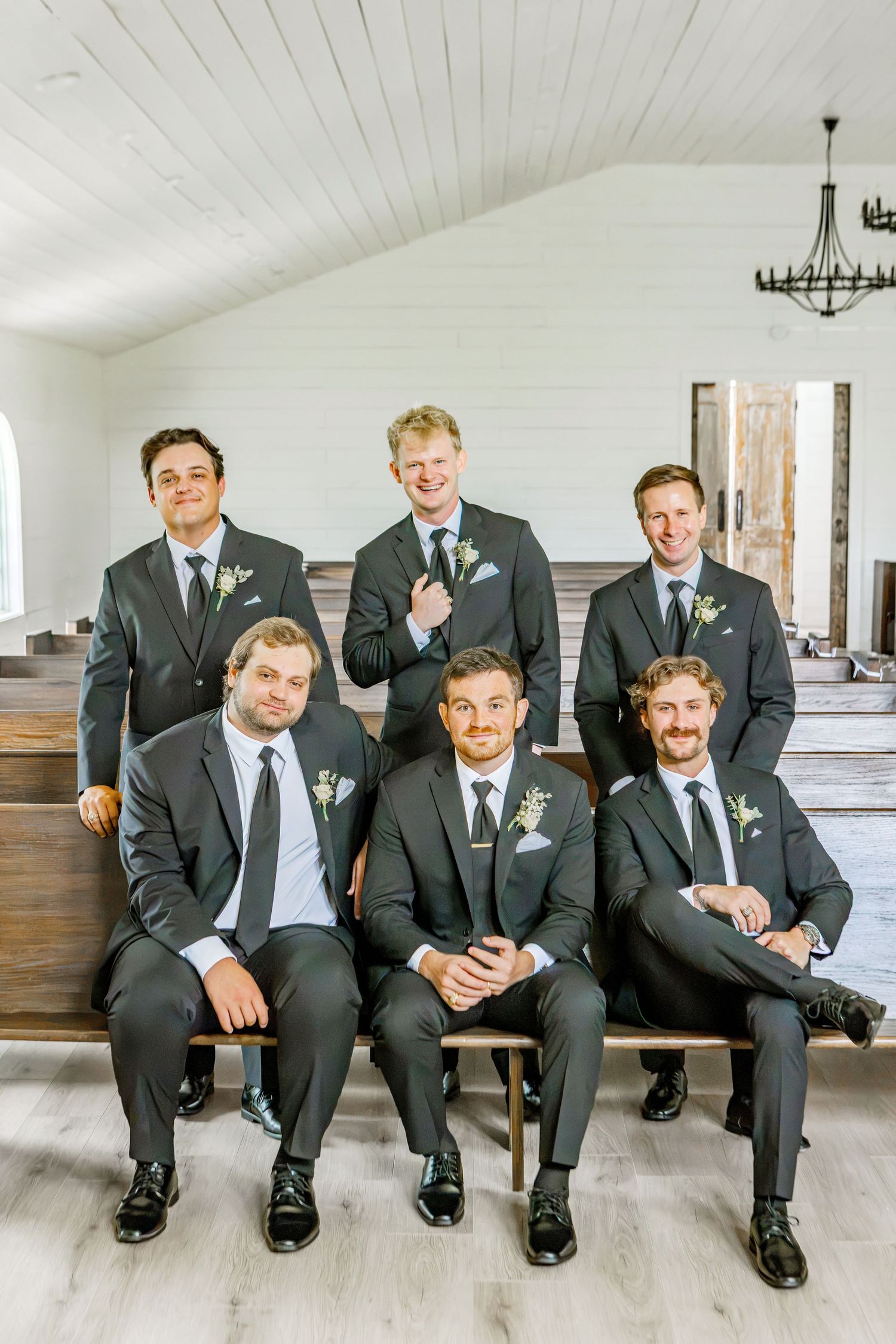Six groomsmen in dark suits with boutonnieres pose in a bright white chapel, three seated on wooden benches and three standing behind.
