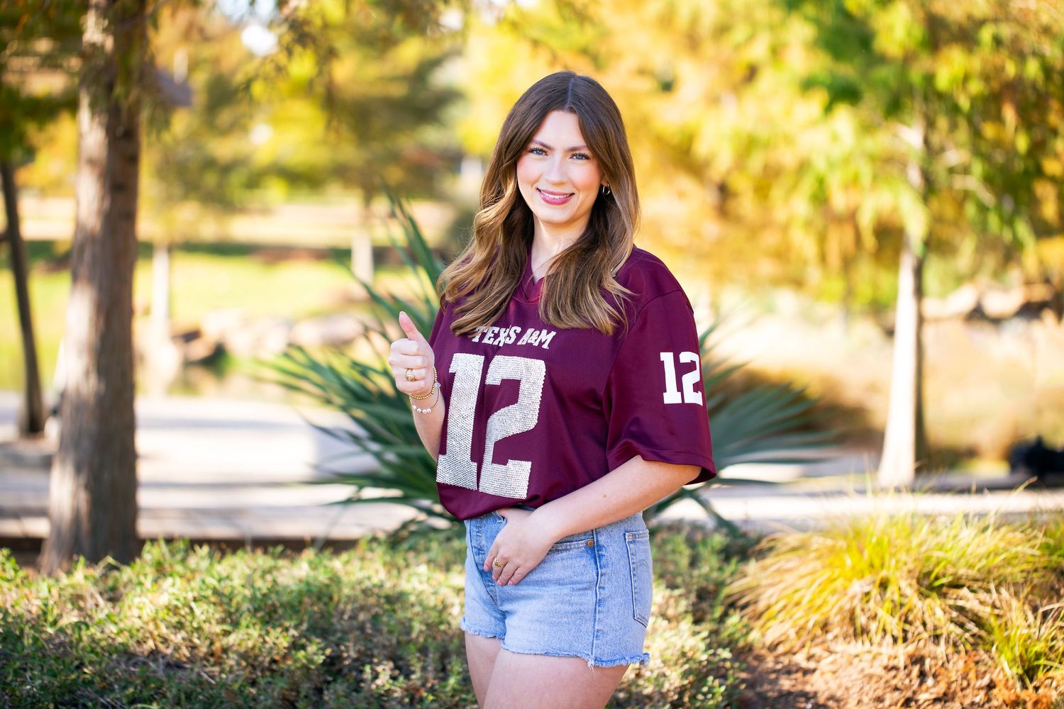 A young woman wearing a purple jersey and shorts stands in a park with trees and greenery in the background.