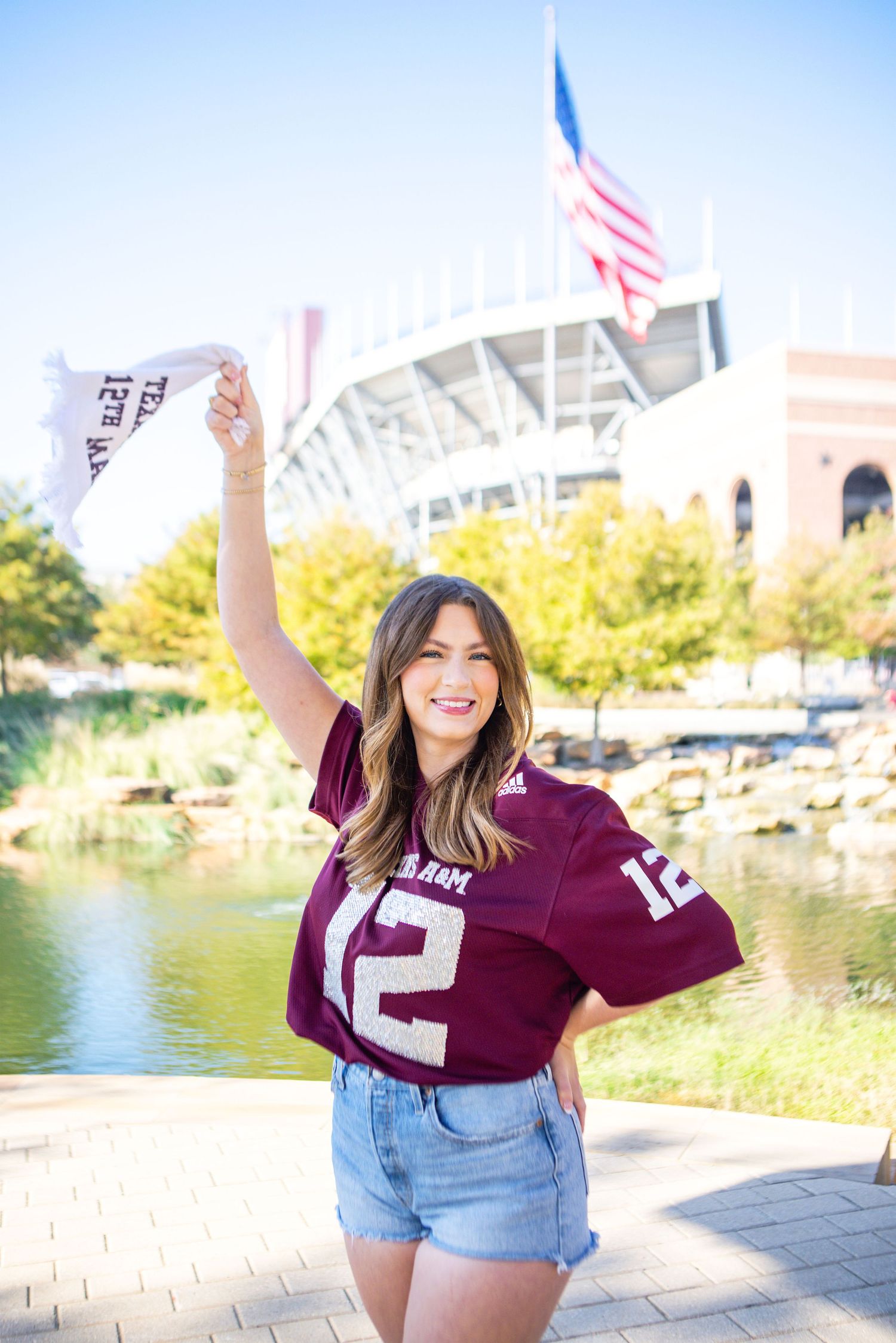 A young woman in a burgundy jersey smiles and waves an American flag in front of a sports stadium.