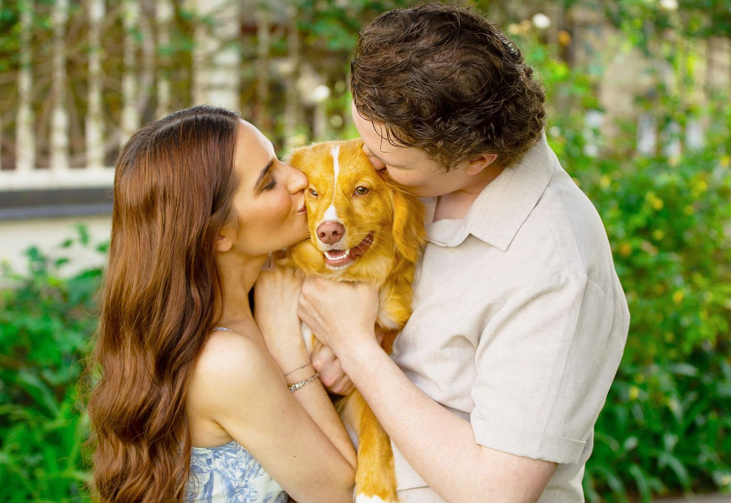 A couple kisses their happy golden retriever dog outdoors in a lush green garden setting.