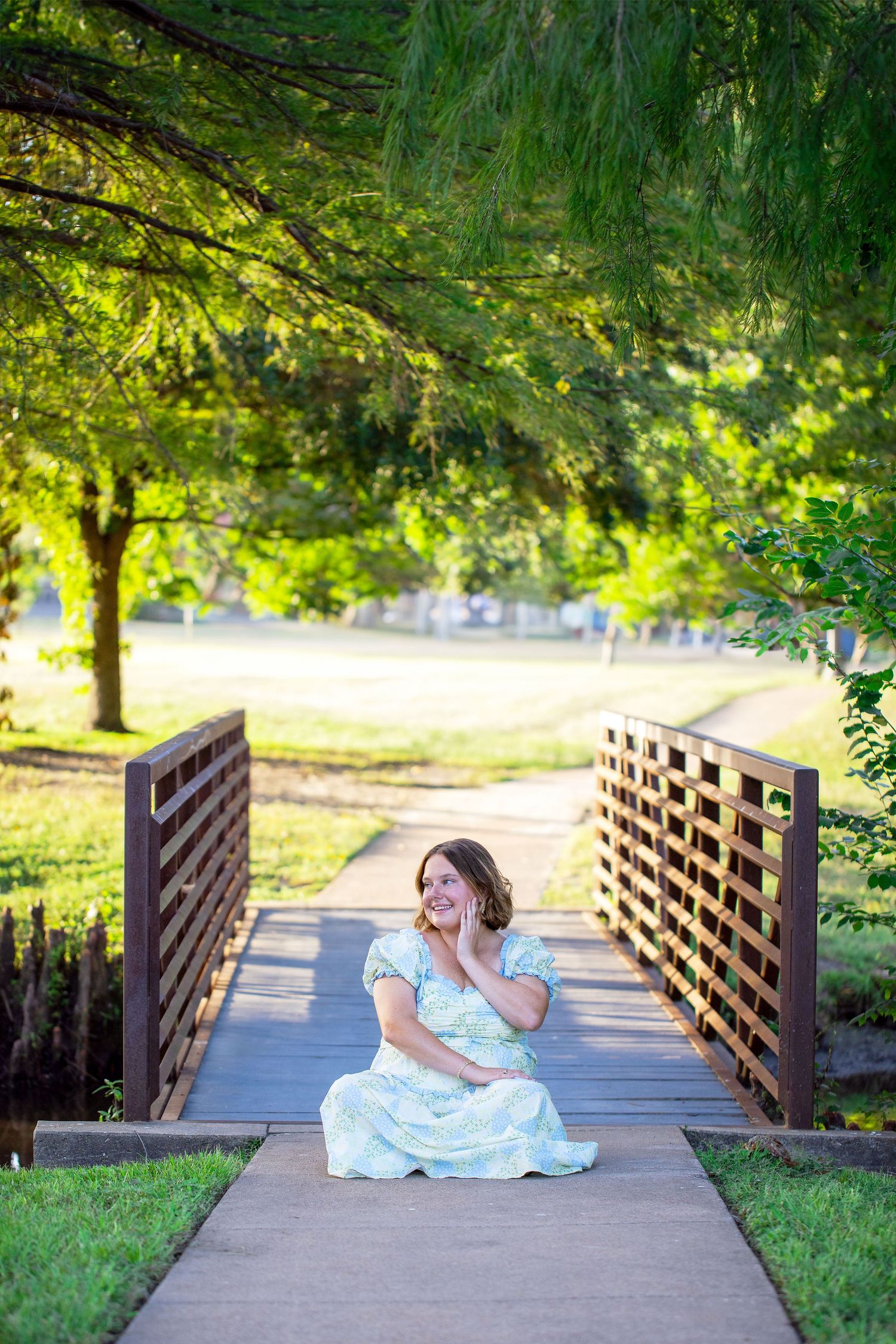 Texas A&M University graduate in light blue dress, photographed on a wooden bridge surrounded by lush green trees at Gabbard Park.