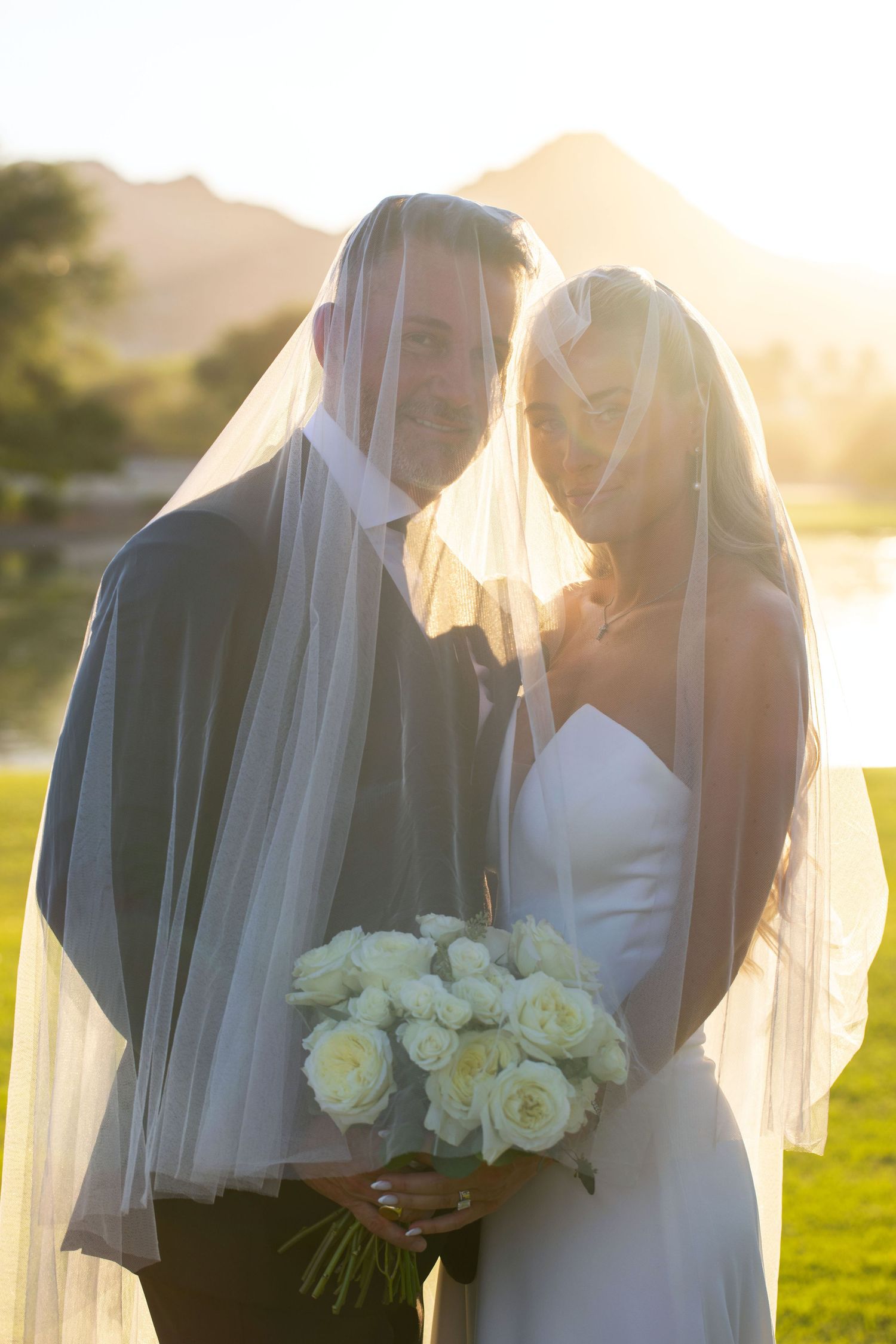 Newlywed couple embraces while holding white rose bouquet in golden sunset light in Scottsdale, Arizona.