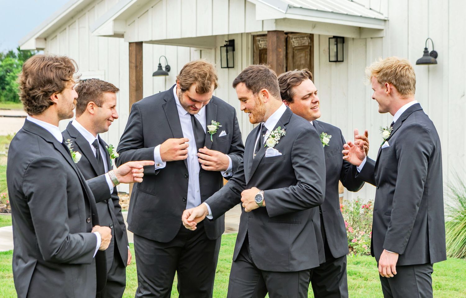 Groomsmen in dark suits laughing and adjusting boutonnieres outside a white barn wedding venue.