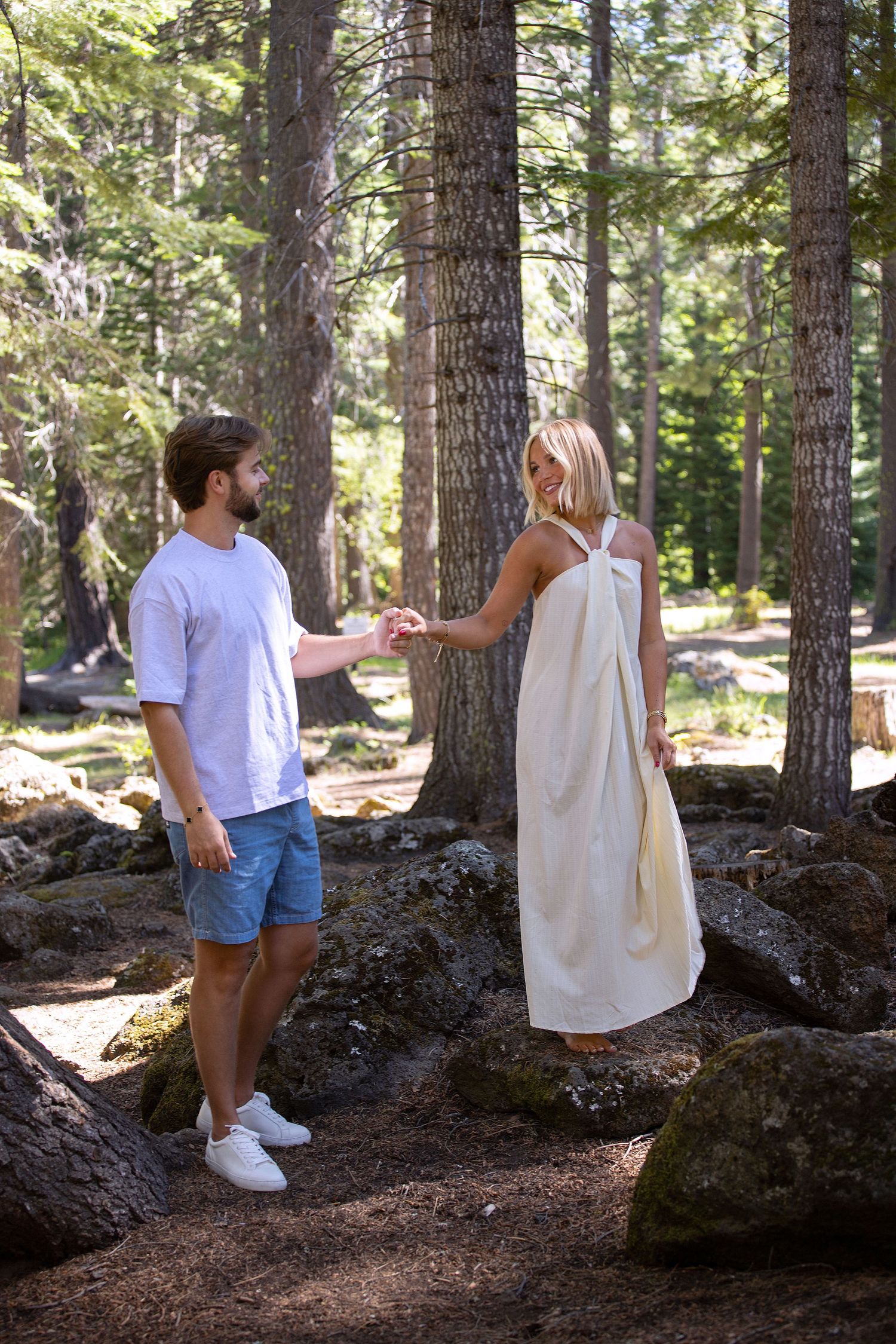 A couple holds hands in a sunlit forest, man in white tee and shorts, woman in elegant cream maxi dress.