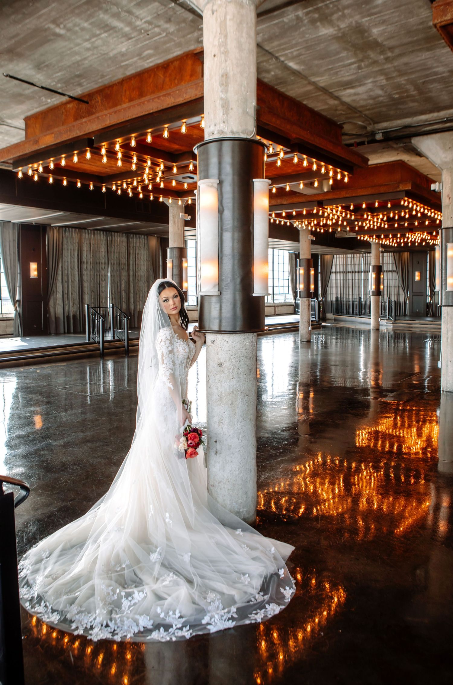 A stunning bridal gown with a flowing train reflects on polished concrete floors in an industrial venue with string lights overhead.