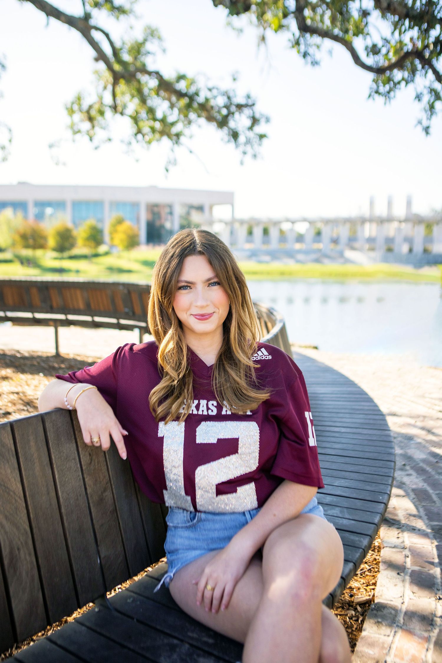 A young woman with long, wavy brown hair wearing a burgundy jersey and sitting on a wooden bench in an outdoor setting.