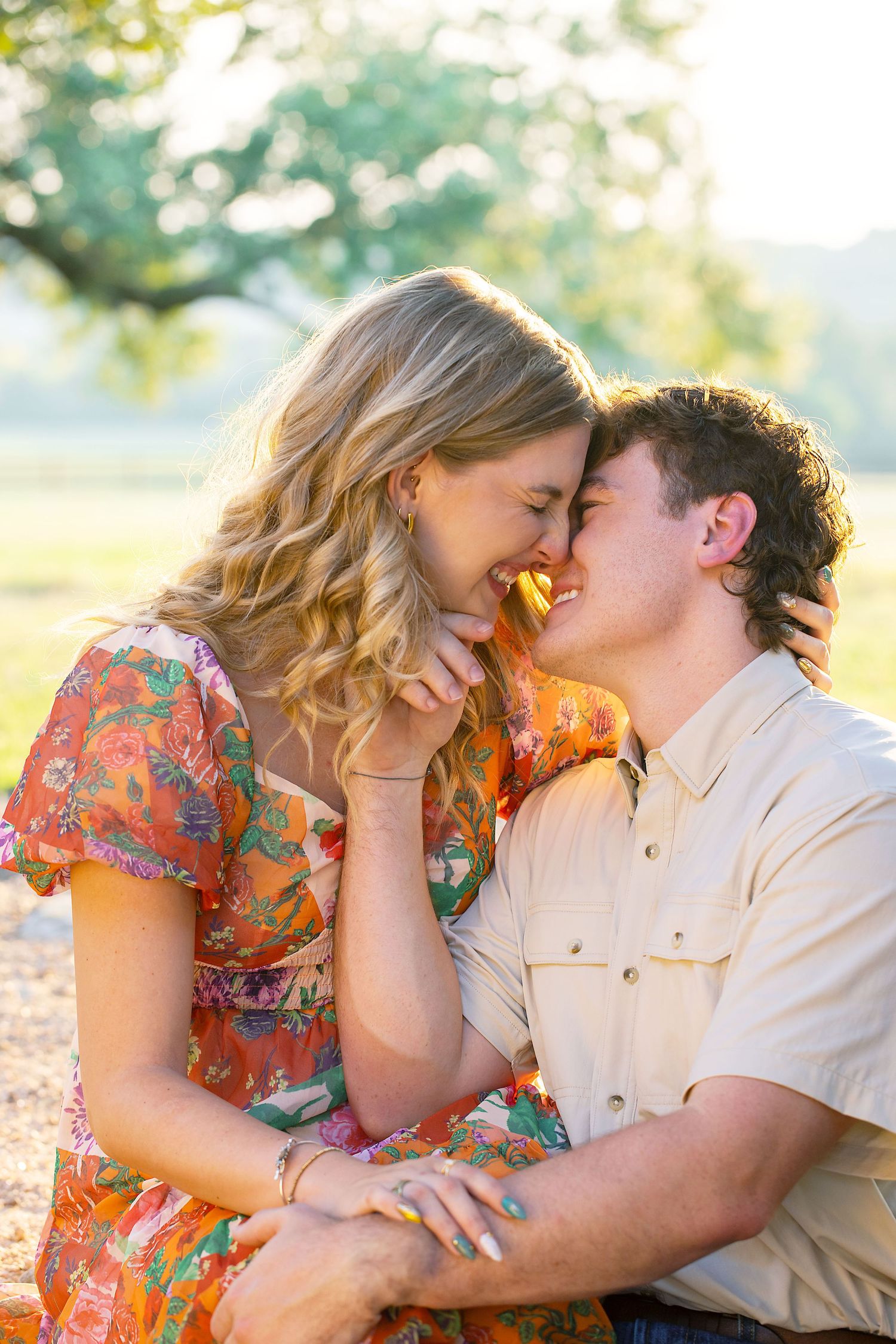 A couple shares an intimate moment during engagement photos in golden sunlight, with her wearing a floral dress and him in a beige shirt.