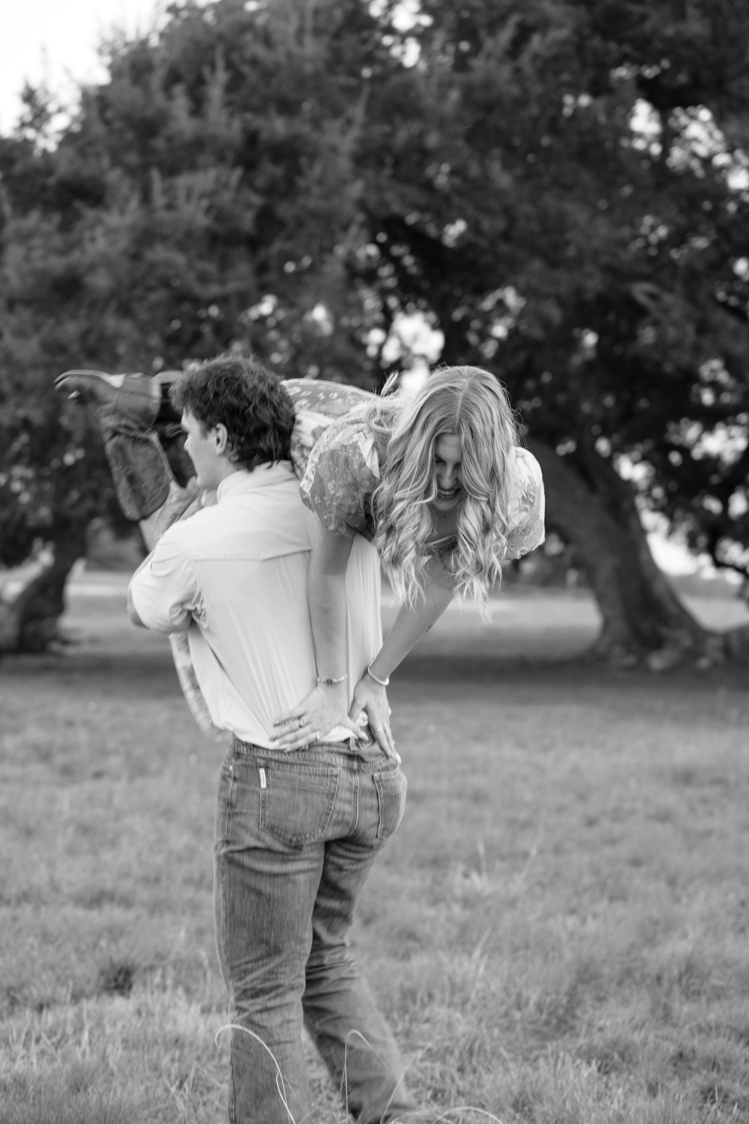 A couple in jeans embraces playfully in a grassy field on a sunny day.