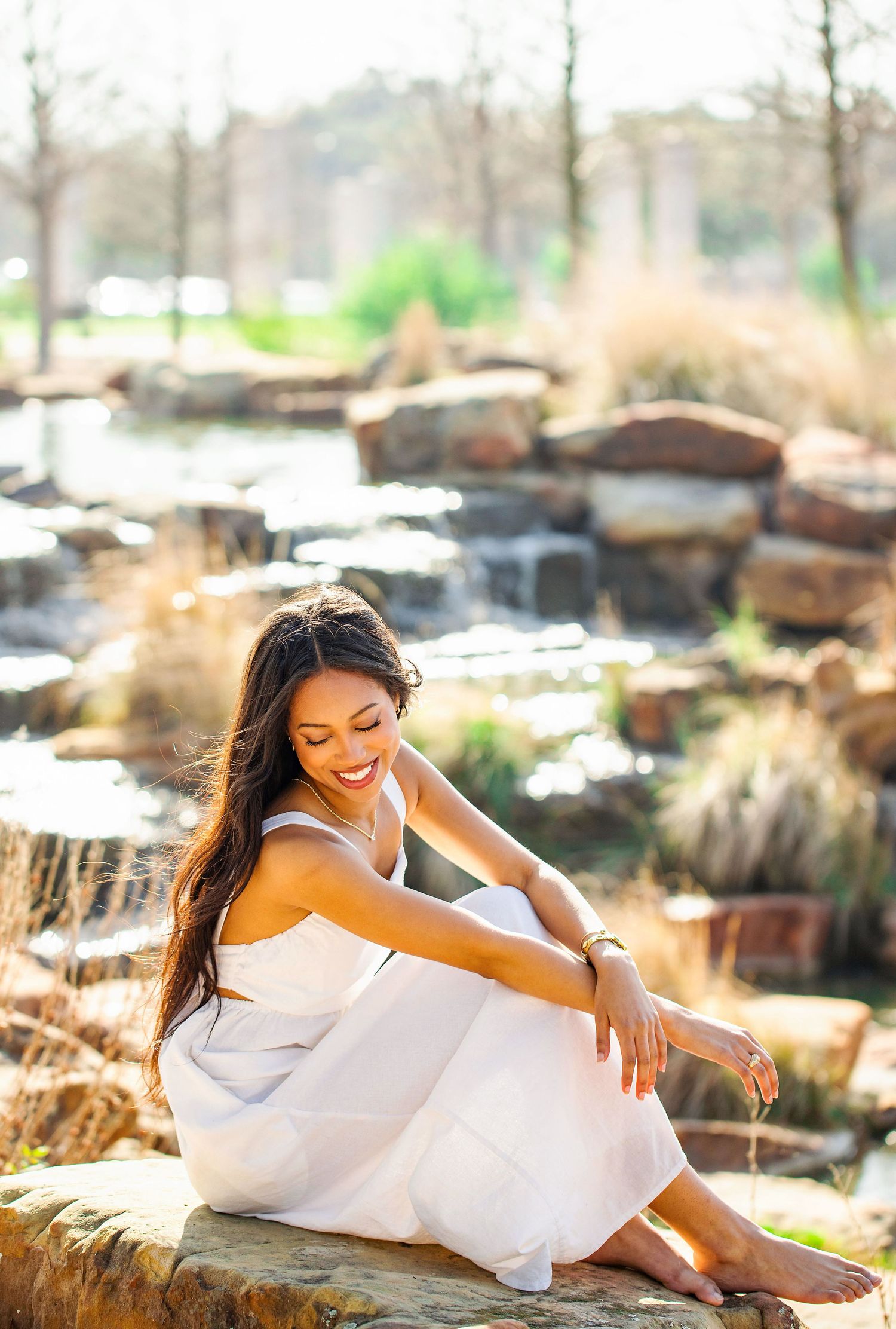 A person in a flowing white dress sits gracefully on stone steps near a waterfall feature in an outdoor garden setting.