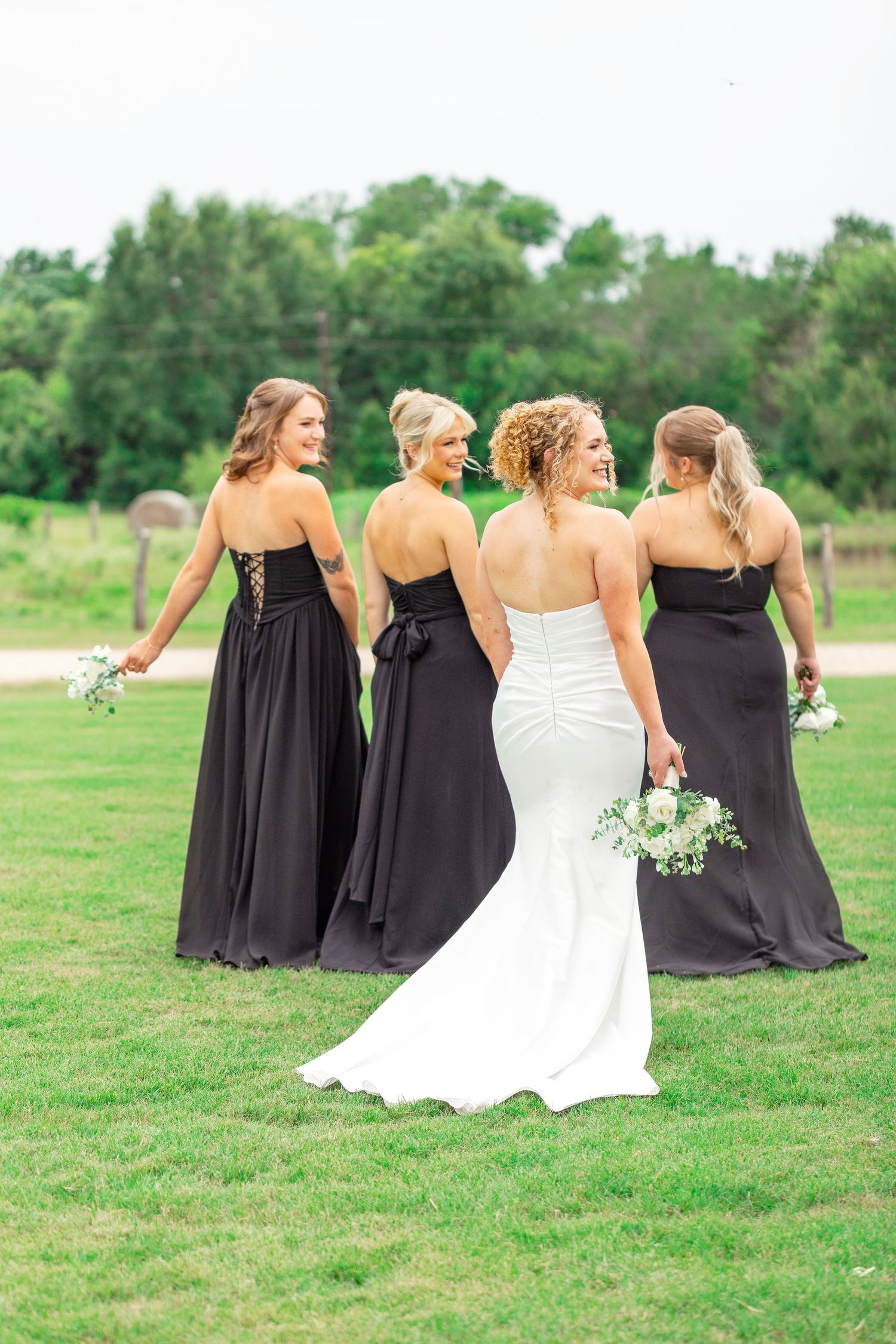 Bride in white gown poses with three bridesmaids in black dresses holding bouquets on green lawn.
