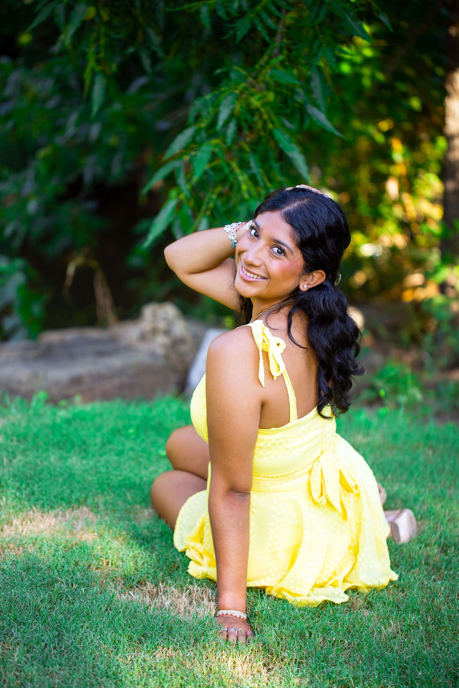 Texas A&M University graduate in yellow sundress poses playfully on green grass during outdoor photo session at the Leach Teaching Gardens.