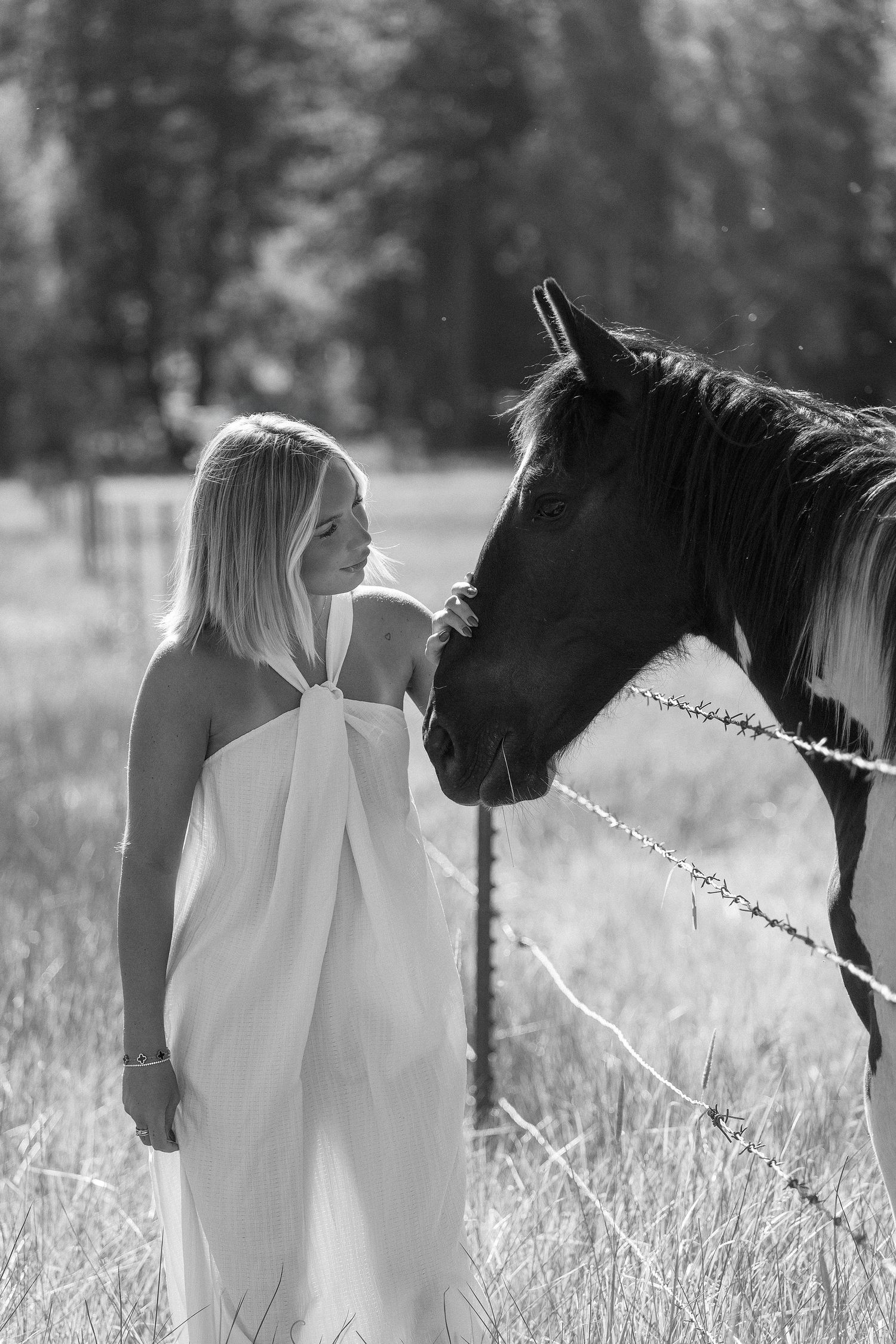 Black and white photo of a blonde woman in a white dress standing beside a horse in a sunny field.