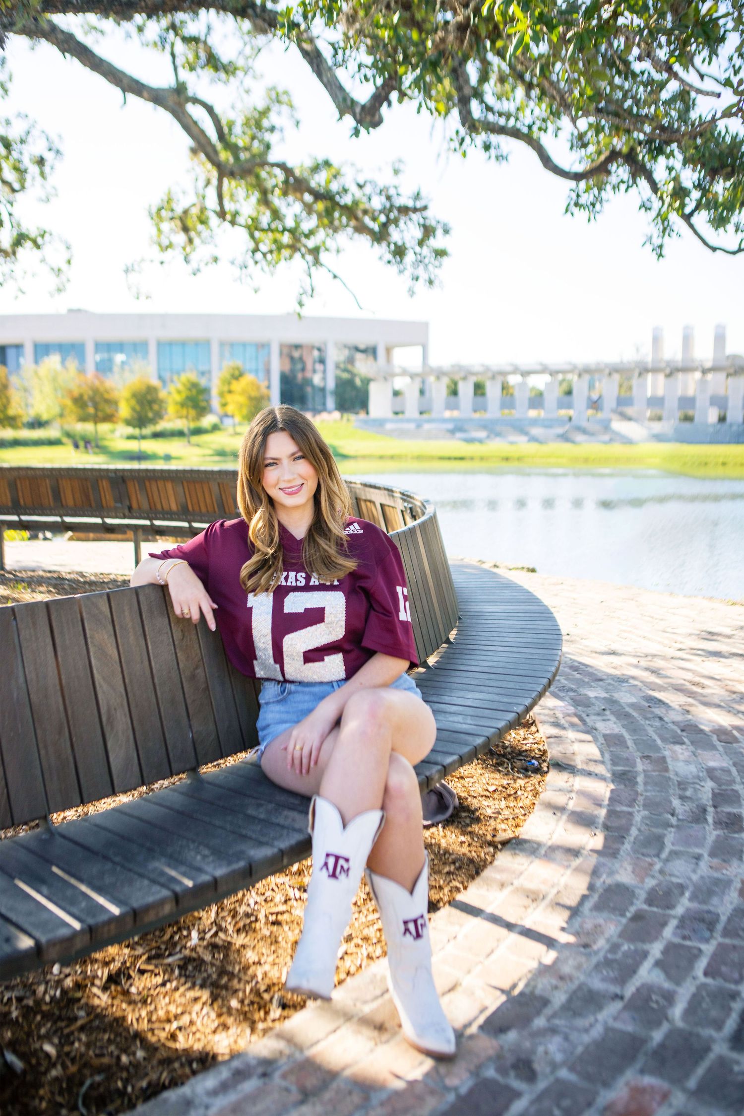 A young woman wearing a jersey with the number 12 sits on a wooden bench near a lake surrounded by trees.