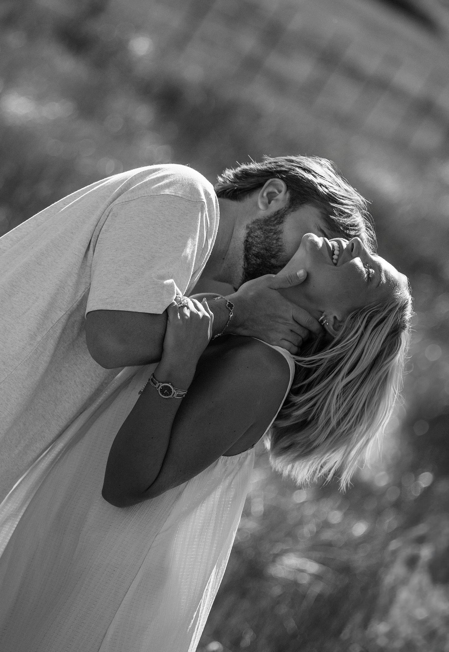 Black and white photo of a couple embracing and kissing outdoors, woman in white dress tilting head back.
