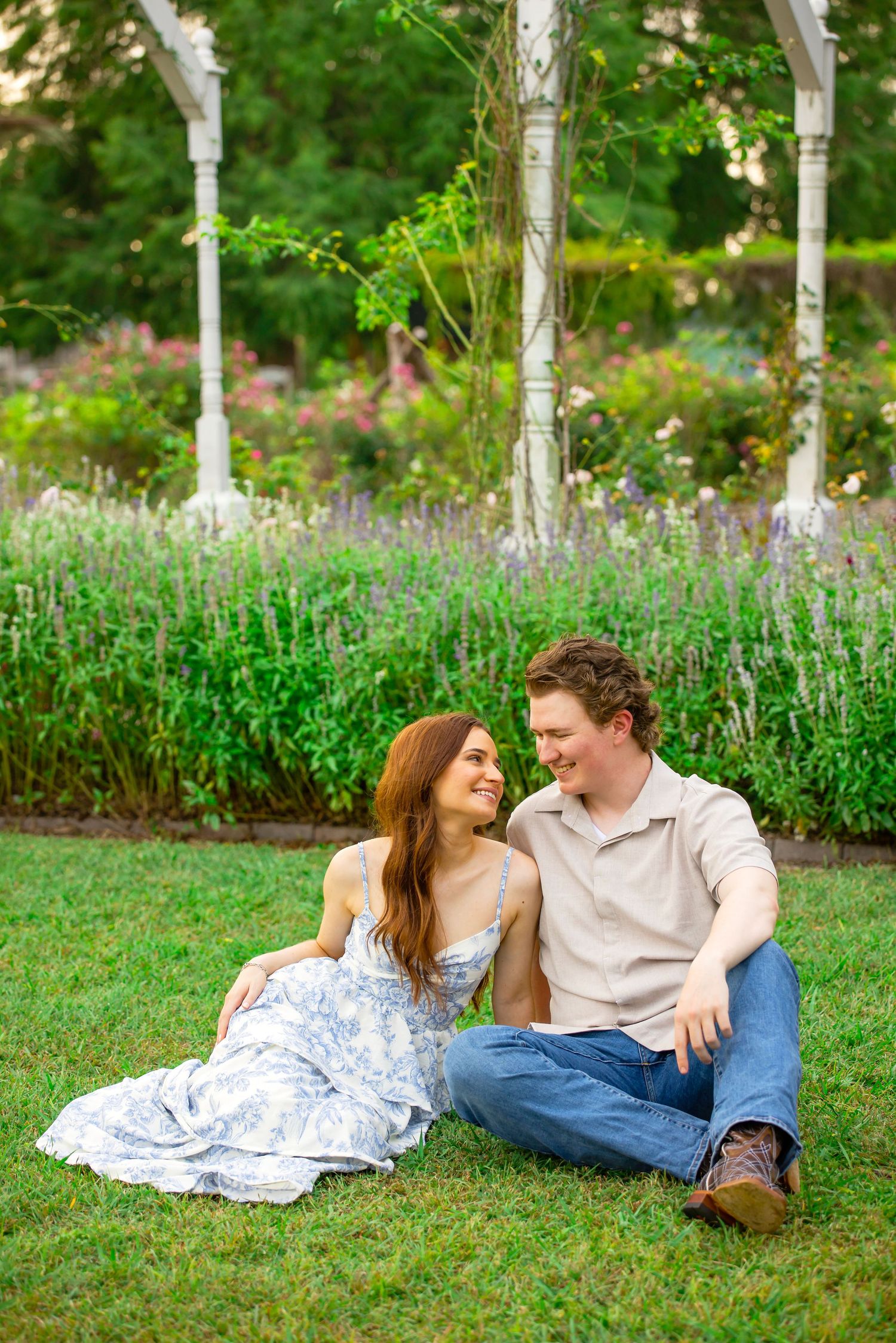 A couple sits together on lush green grass in a garden, smiling at each other surrounded by colorful blooming flowers.