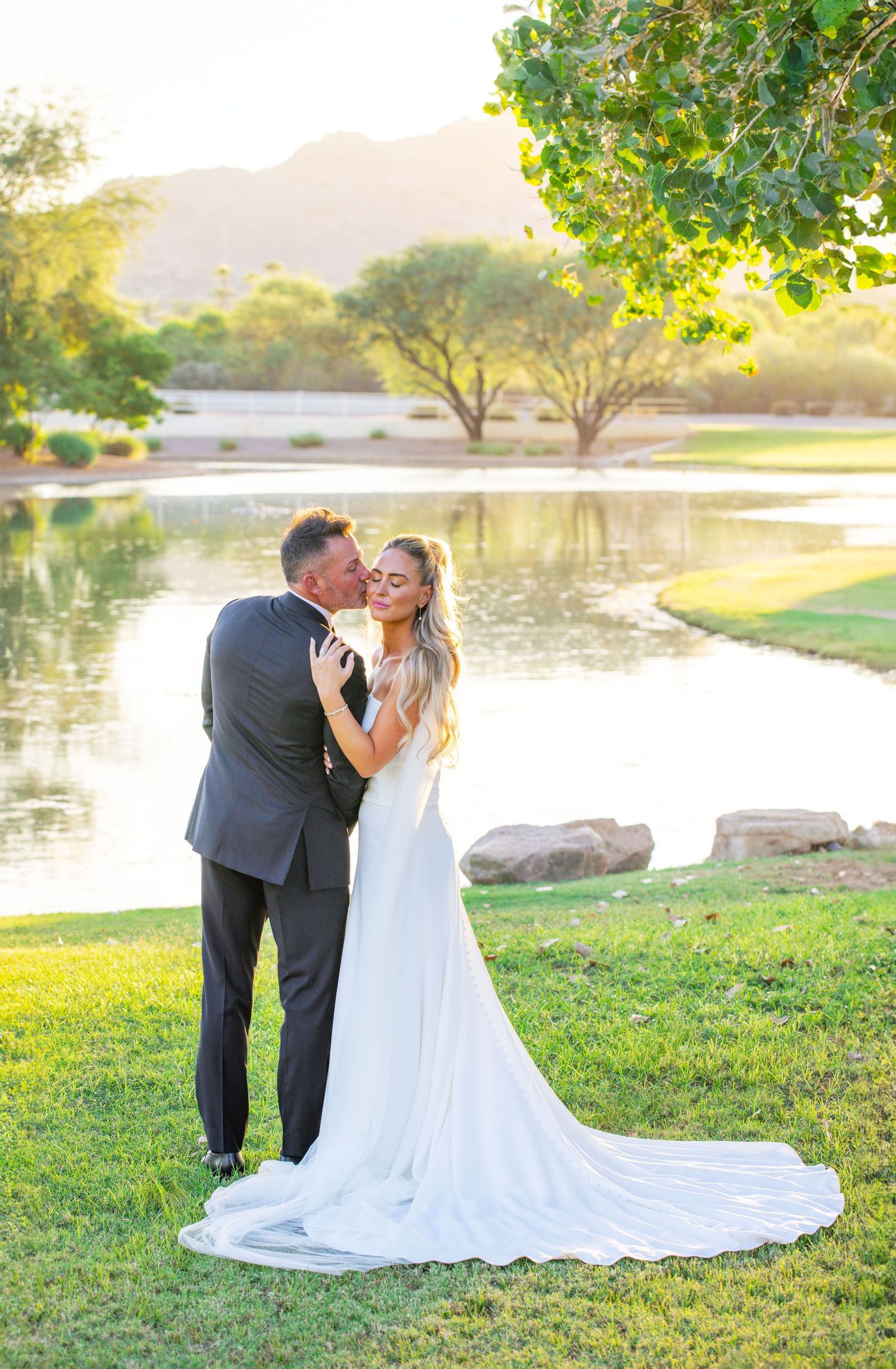 Newlyweds kiss by a serene lake at sunset with mountains, with the bride's flowing white dress trailing behind in Scottsdale, Arizona.