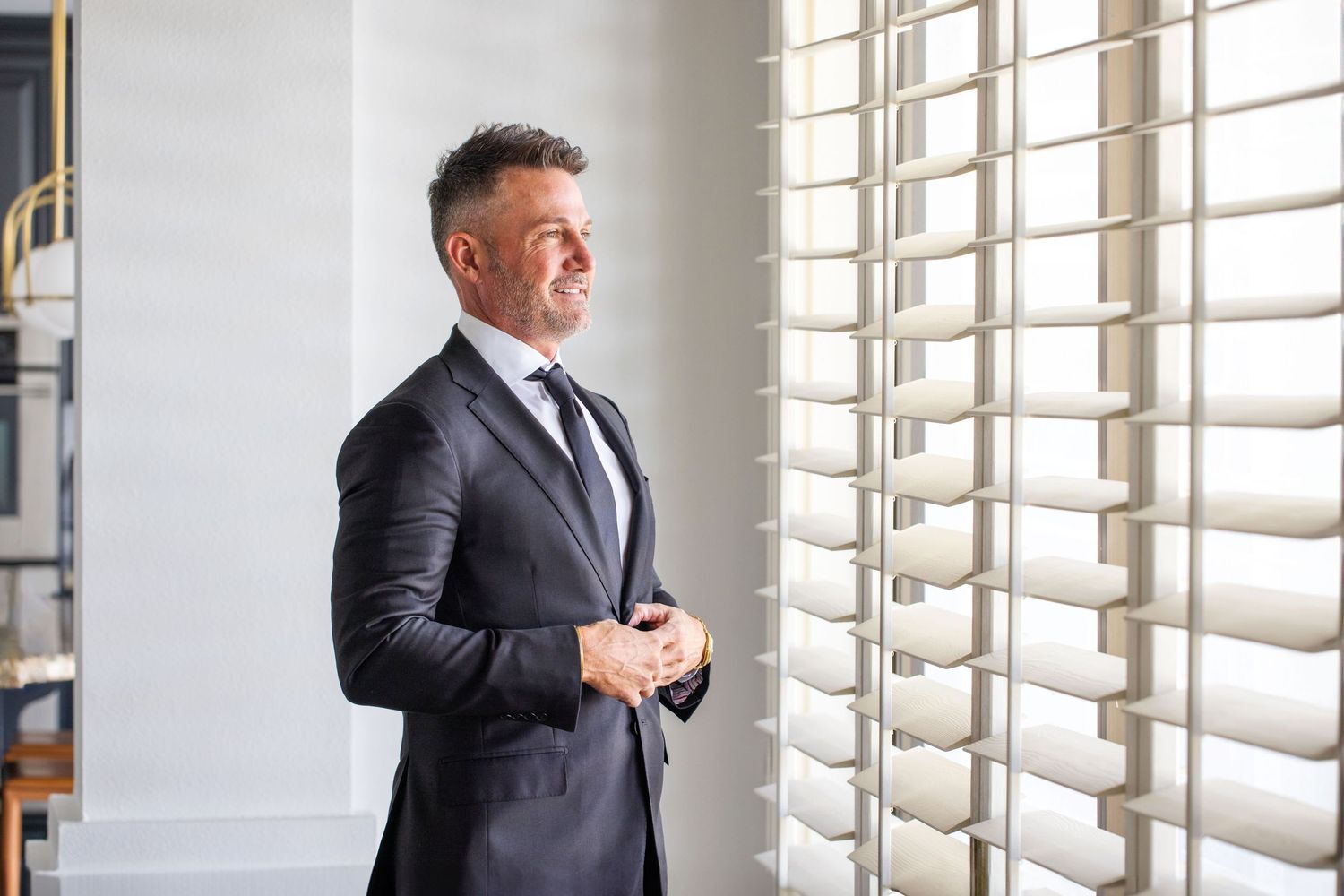 Groom in dark suit adjusts tie near window with natural lighting in Scottsdale, Arizona.