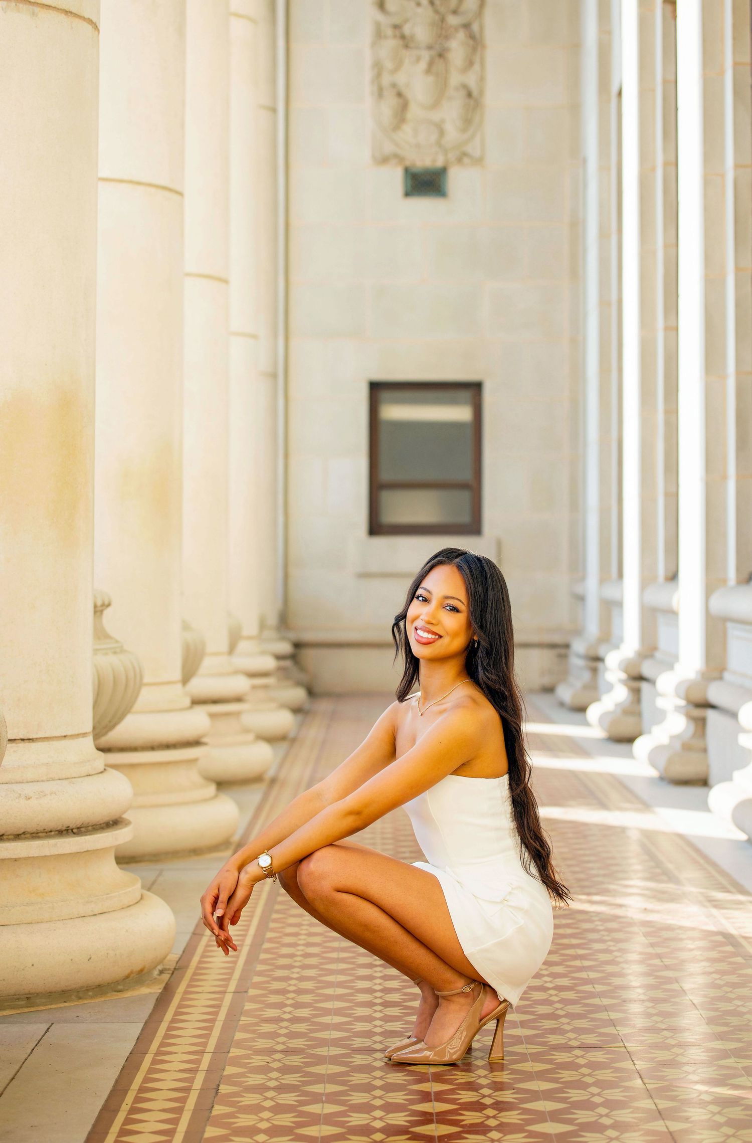 Graduate in white dress poses elegantly on floor between ornate marble columns during photoshoot.