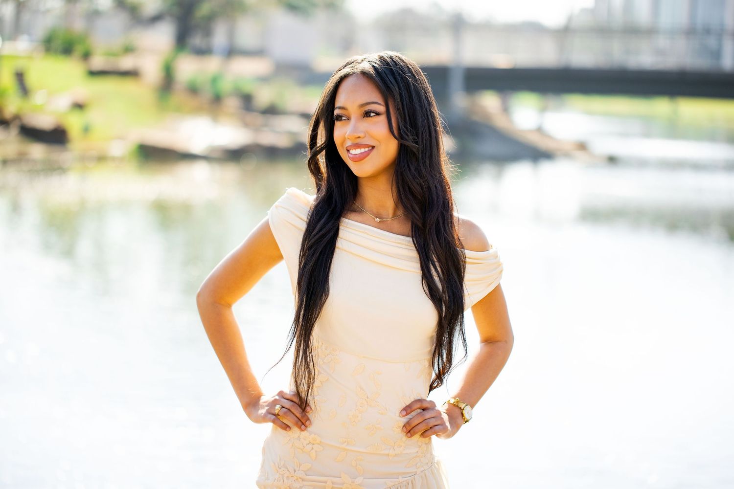 A person in a white dress poses outdoors near a lake or pond on a sunny day.