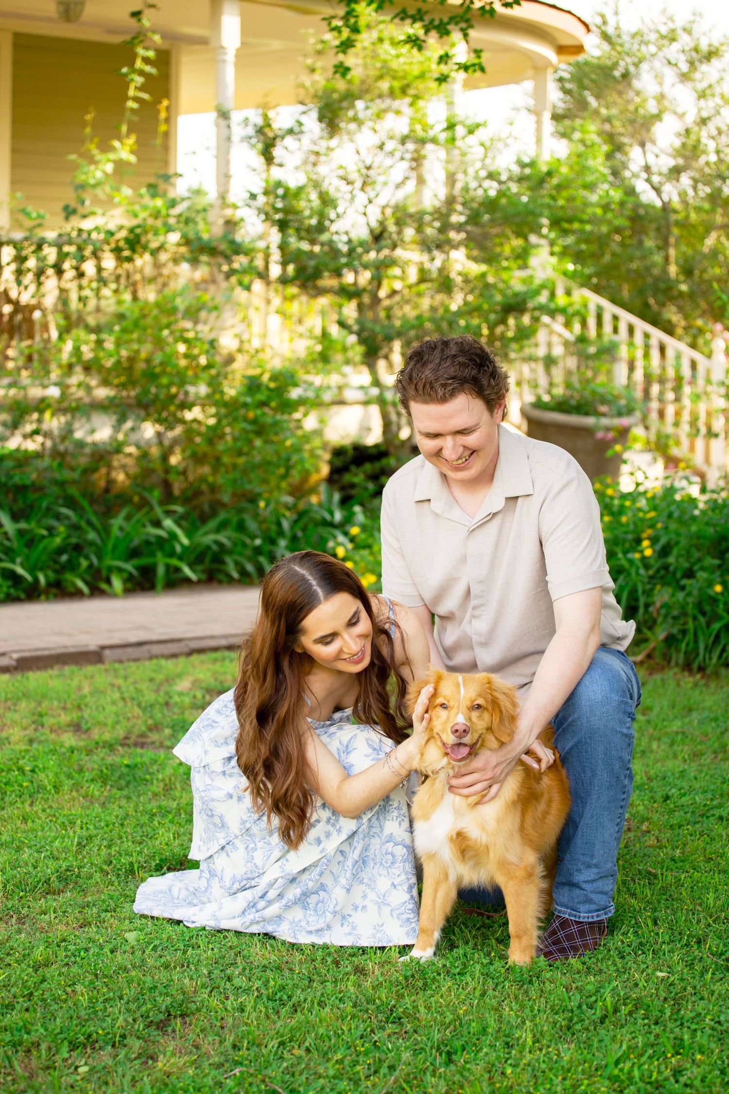 A smiling couple sits on green grass petting their golden retriever dog in front of a charming yellow house.