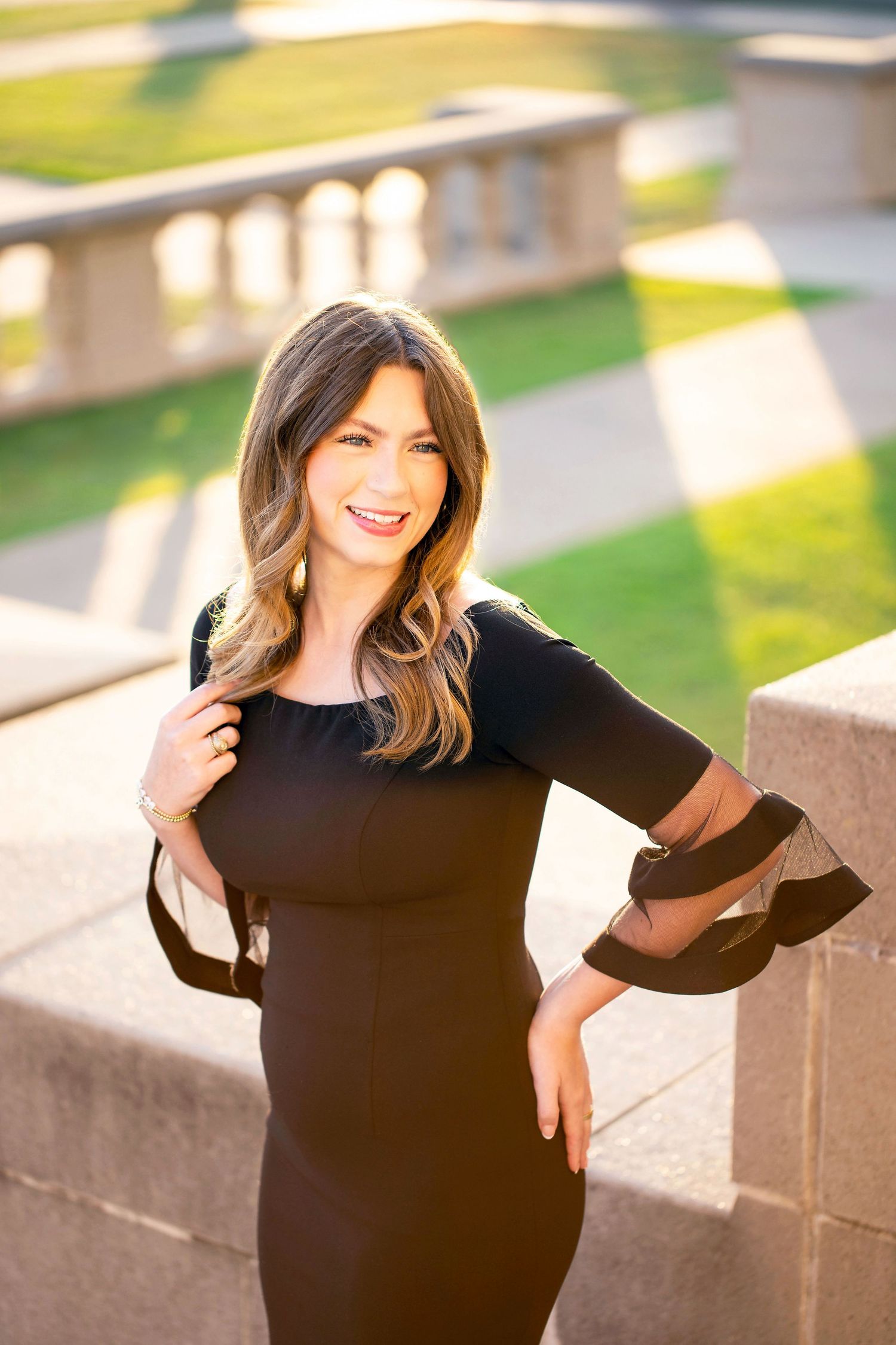 A Texas A&M University graduate in a black dress poses elegantly against a stone wall at the Admin Building in warm sunlight.