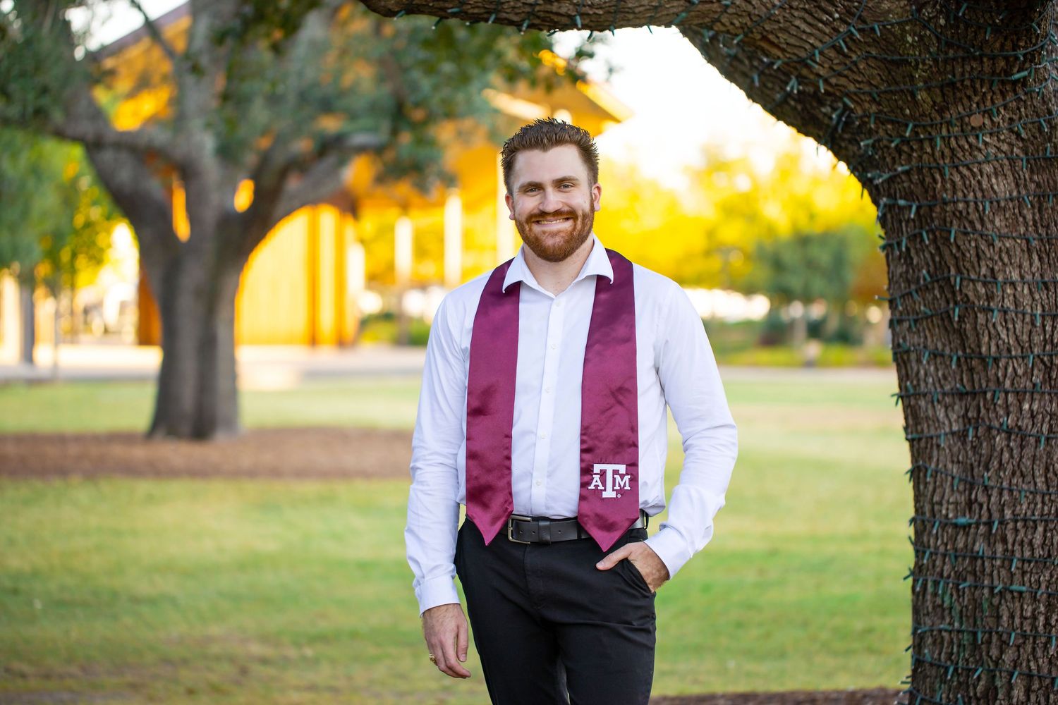 A Texas A&M University graduate in formal attire with a maroon tie smiling outdoors at Aggie Park.