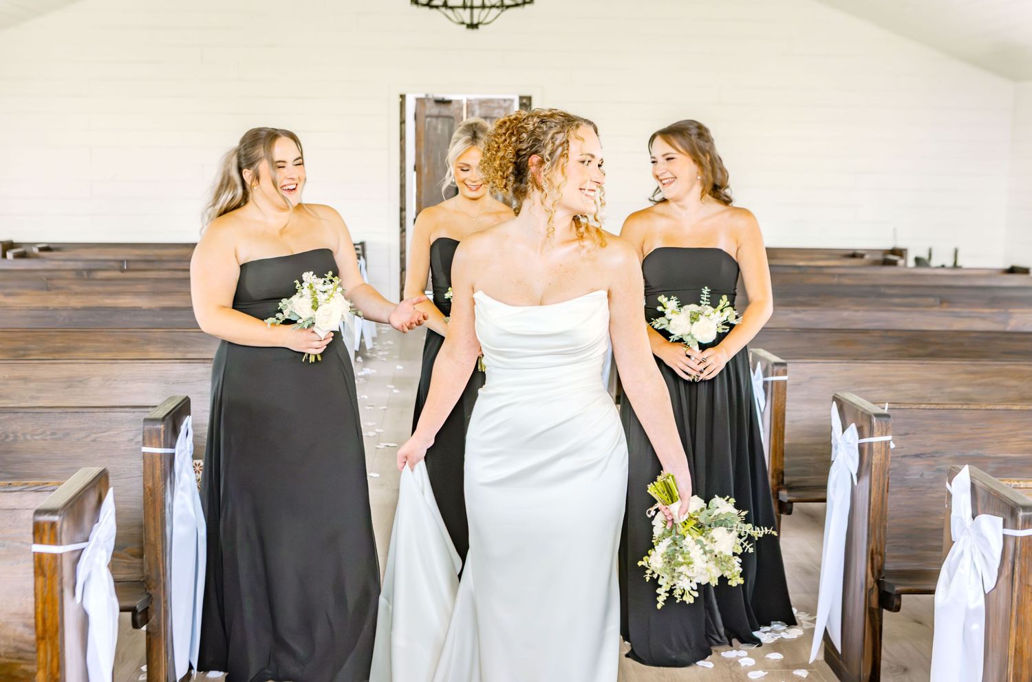 Bride in white strapless gown walks with three bridesmaids in dark dresses down church aisle holding bouquets.