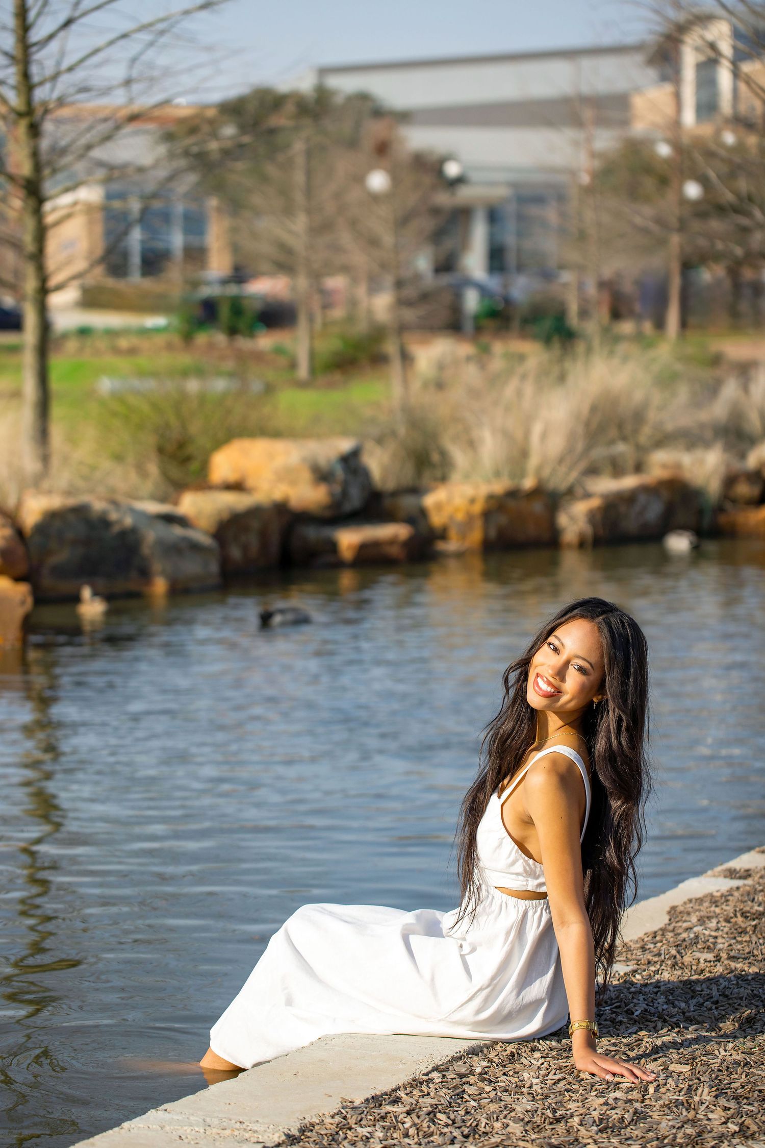 Graduate in flowing white dress poses by waterfront with trees and buildings in background.