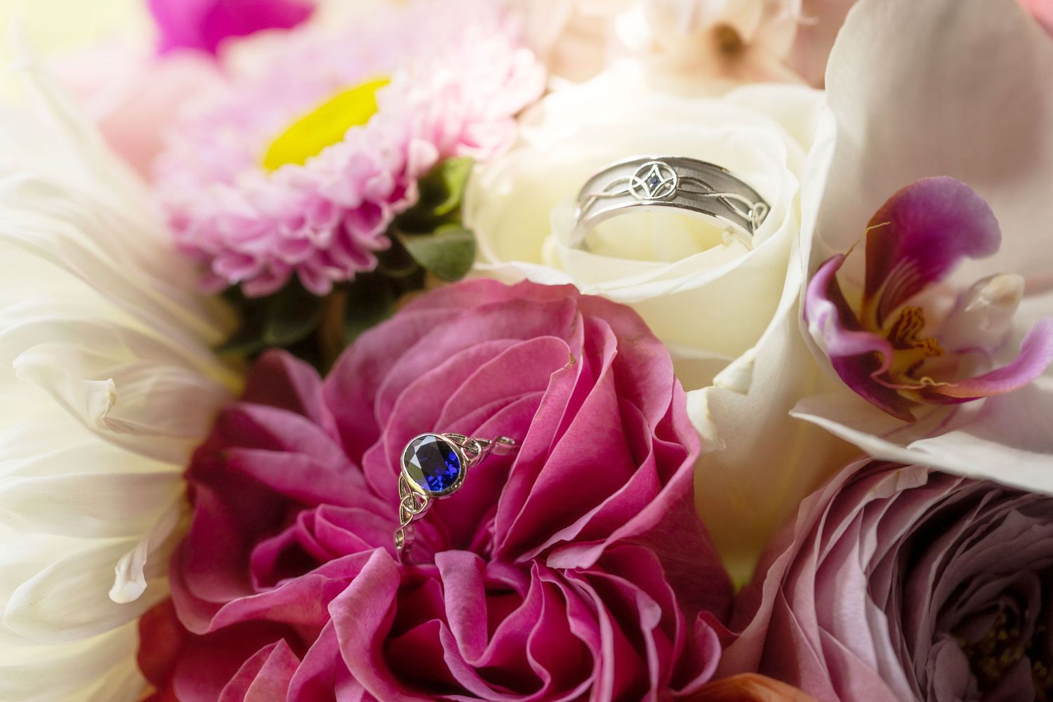 Two elegant rings rest among vibrant pink peonies and white orchids in a romantic bridal bouquet arrangement.