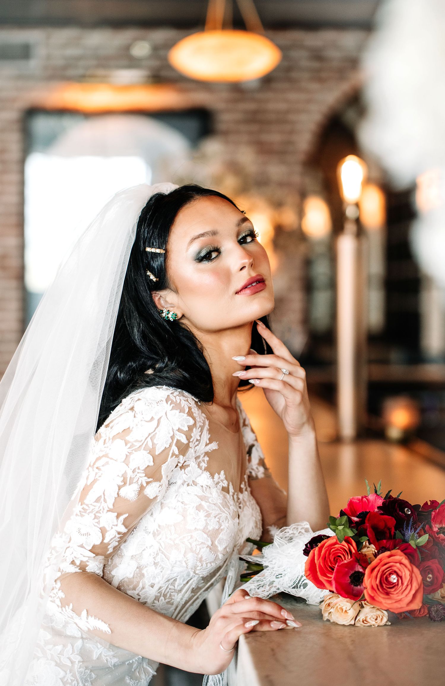 A bride in a delicate lace wedding dress poses thoughtfully with red roses in an elegant indoor venue.