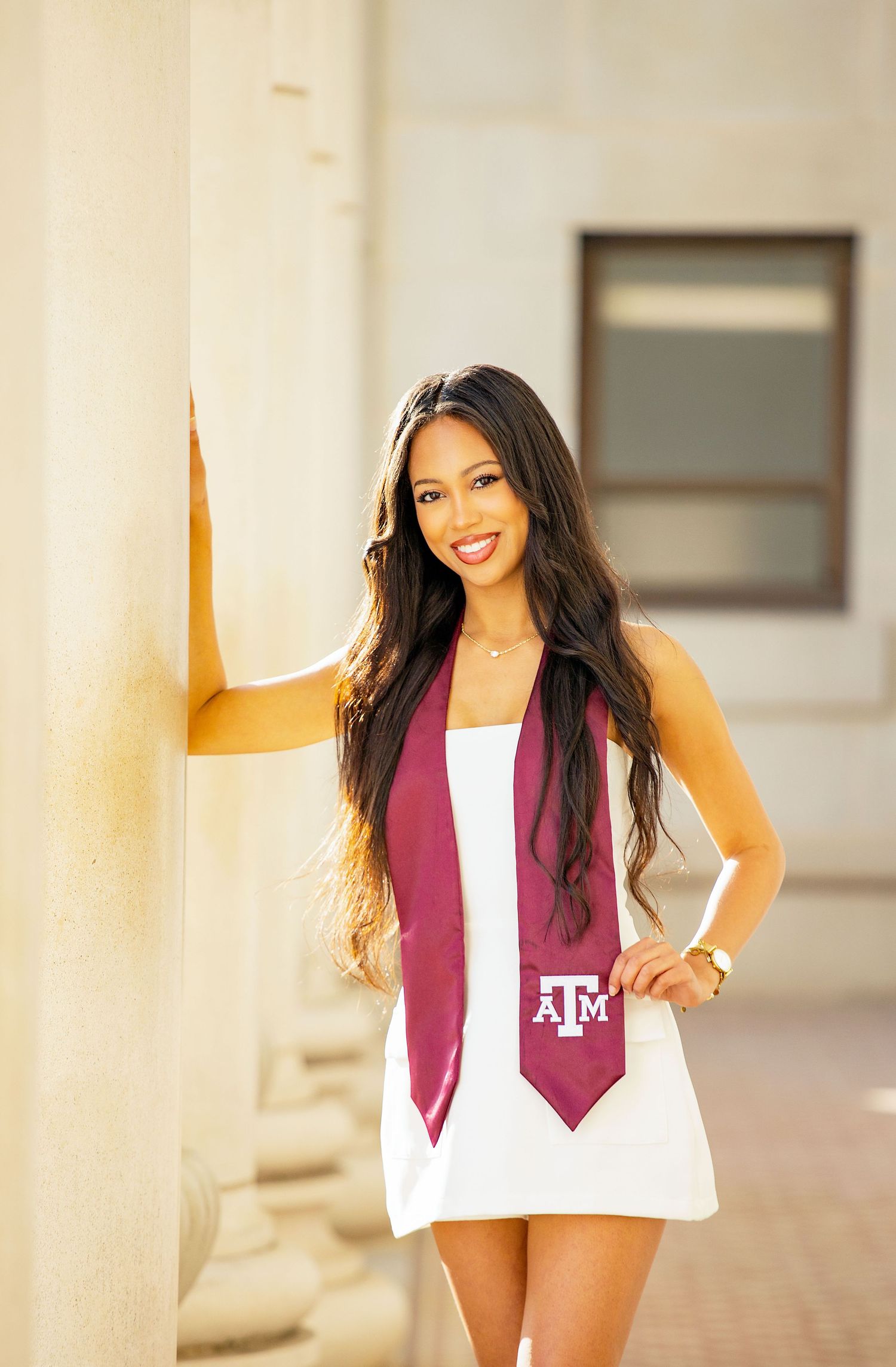 Graduate wearing maroon and white stole with Texas A&M logo poses in front of white columns during graduation photoshoot.