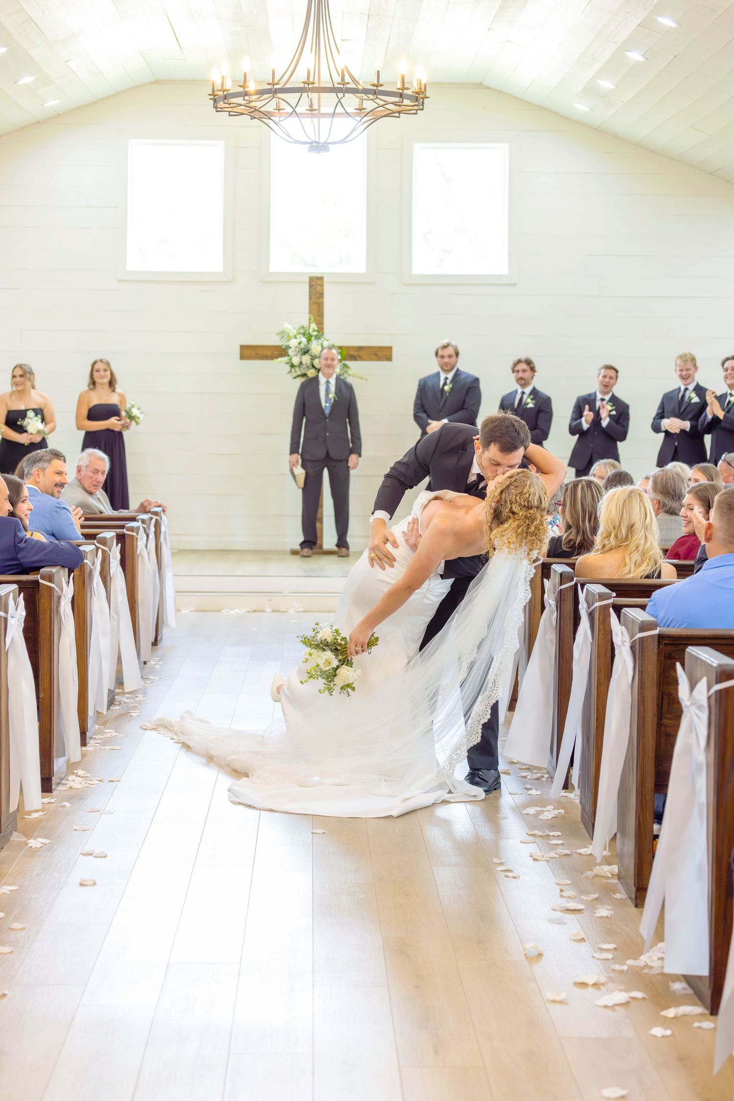 Groom dips bride in wedding chapel aisle after ceremony, guests watching from wooden pews, cross and wedding party behind them.