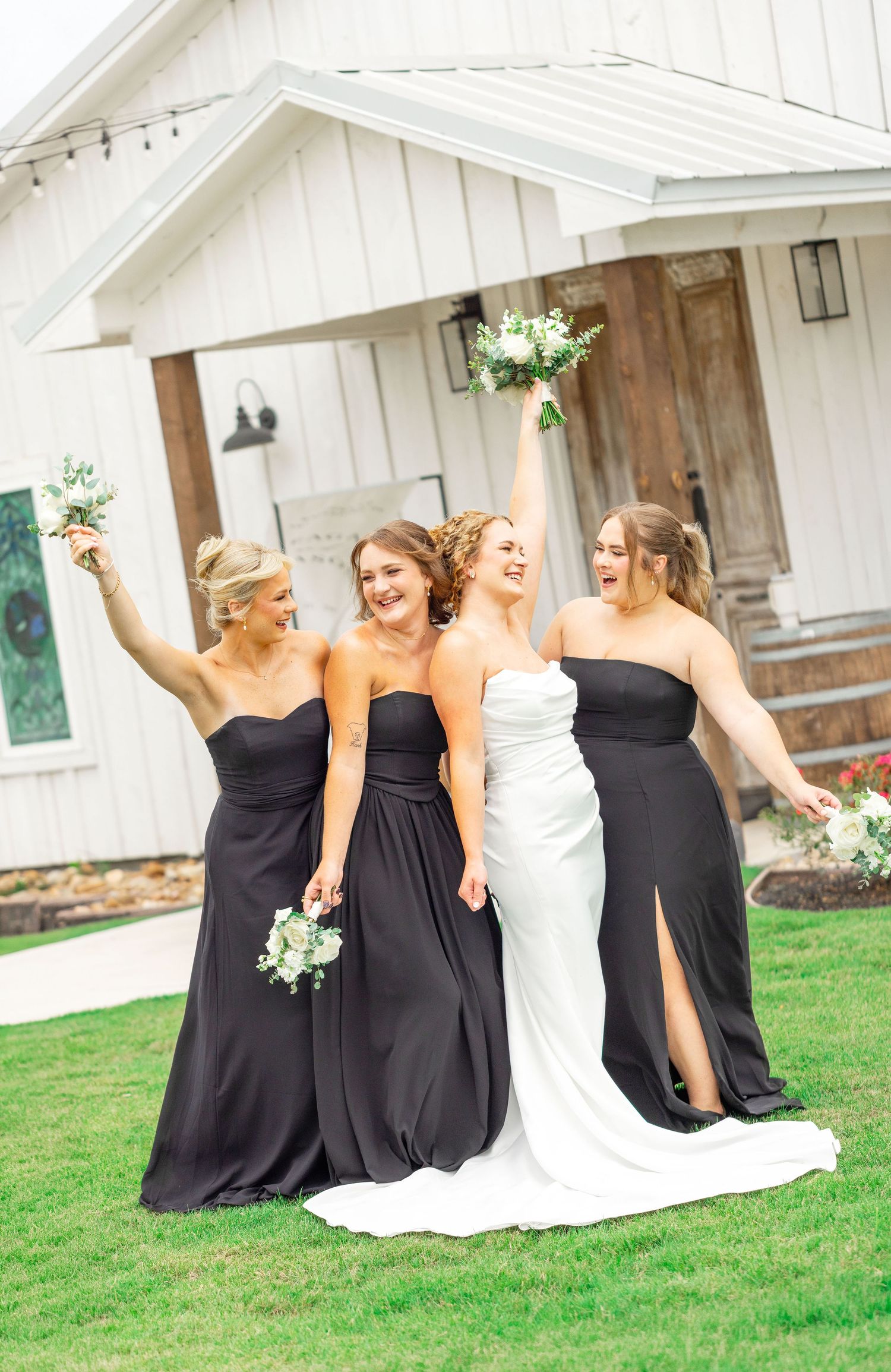 Bride in white gown with three bridesmaids in black dresses holding bouquets aloft outside a white barn venue.