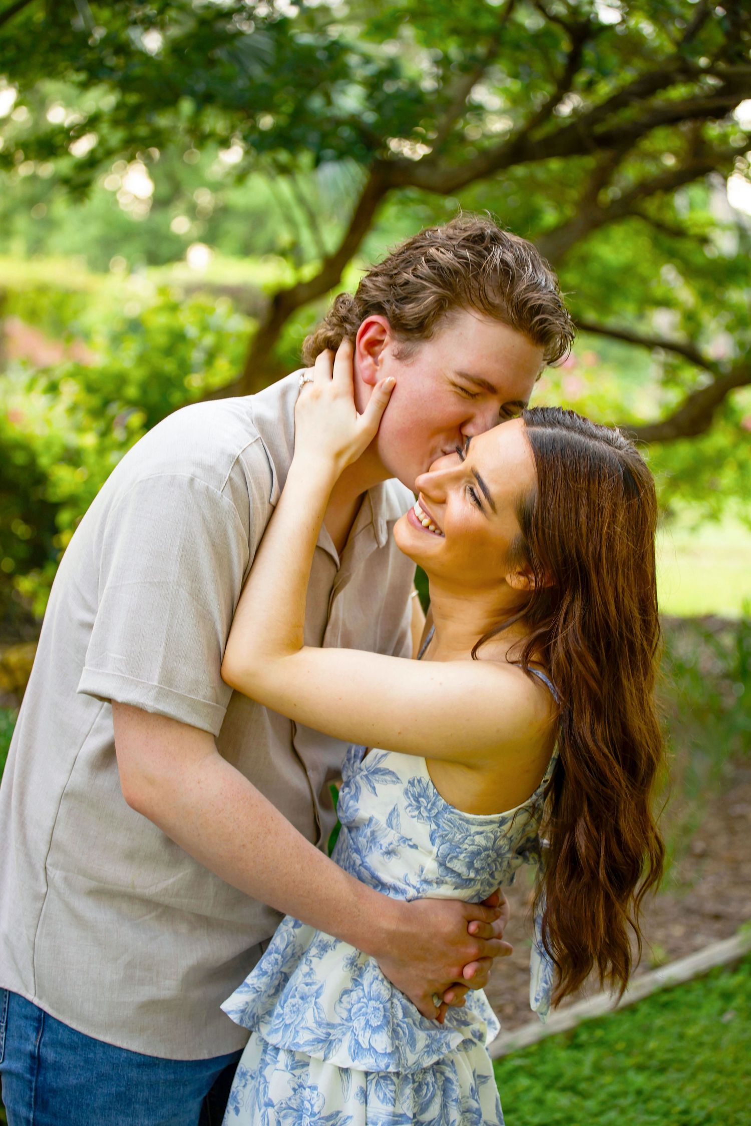 Couple sharing a romantic kiss outdoors, surrounded by lush green trees in a garden setting.