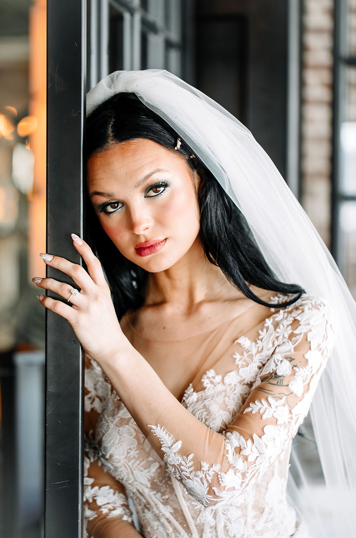 Beautiful bride in white lace wedding dress with delicate floral details and long veil poses in romantic lighting.
