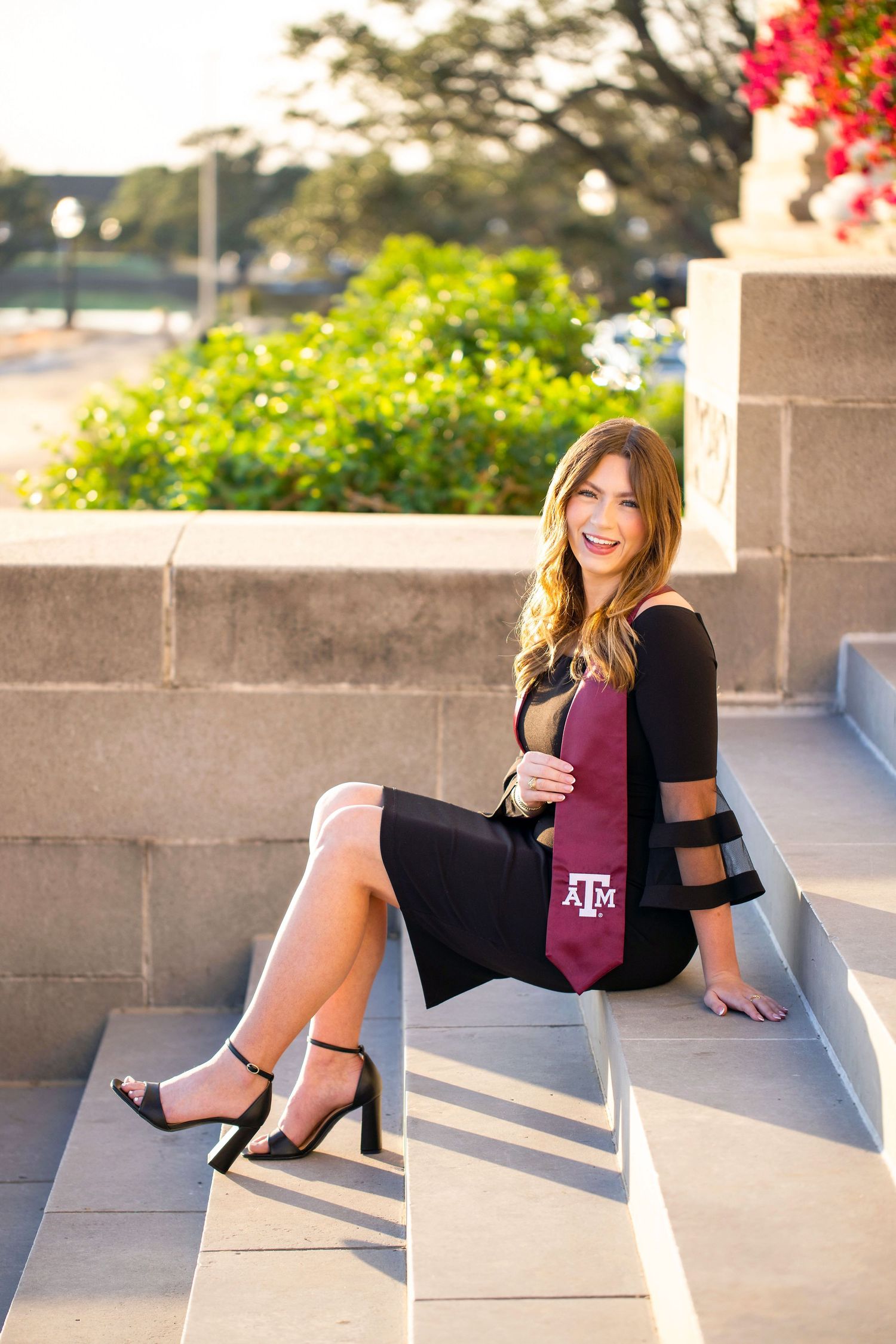 A young woman with long brown hair is sitting on concrete steps, smiling and wearing a black top and pink skirt.