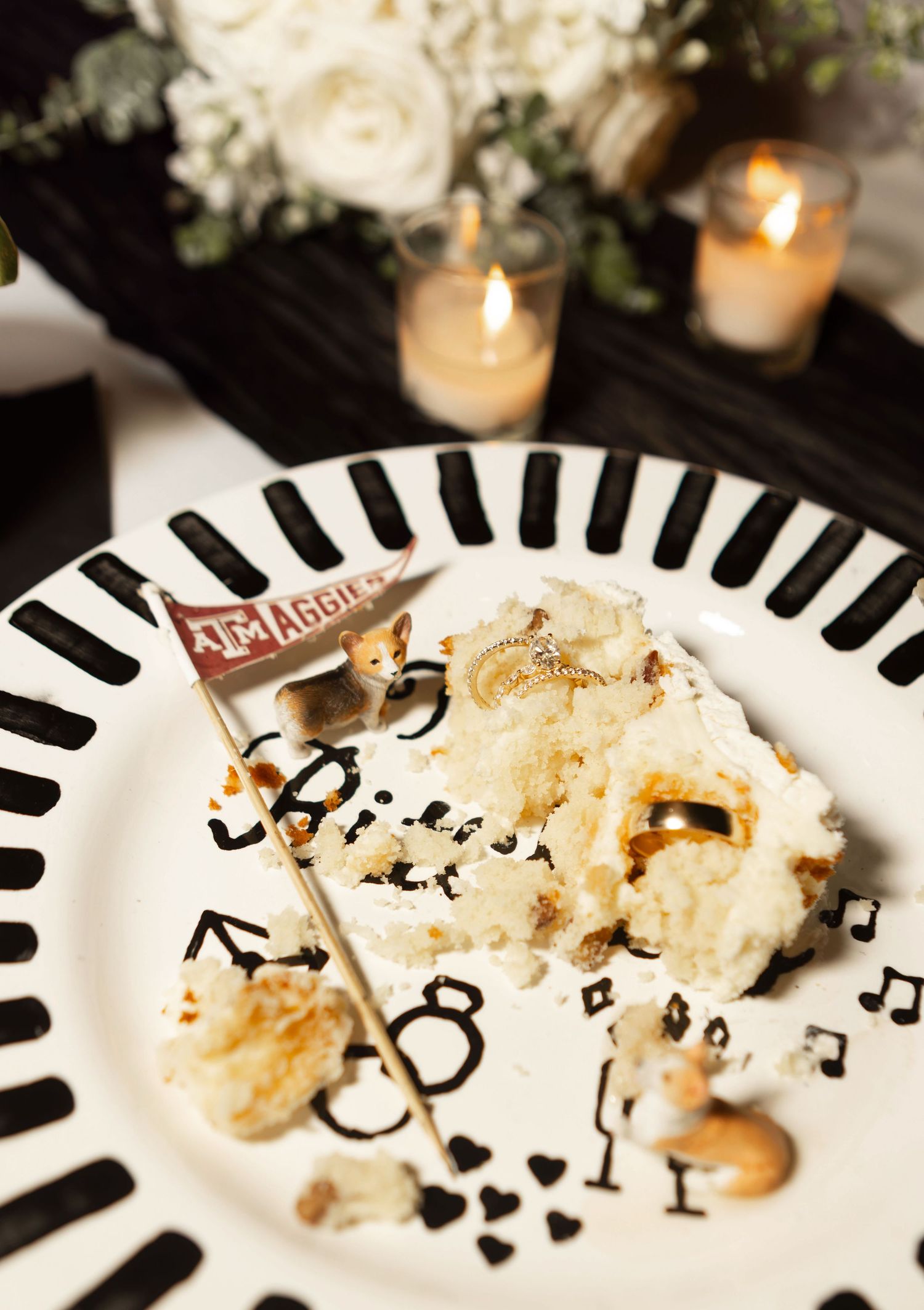 A slice of white wedding cake on a clock-themed plate with an Texas A&M Aggies pennant topper, candles in background.