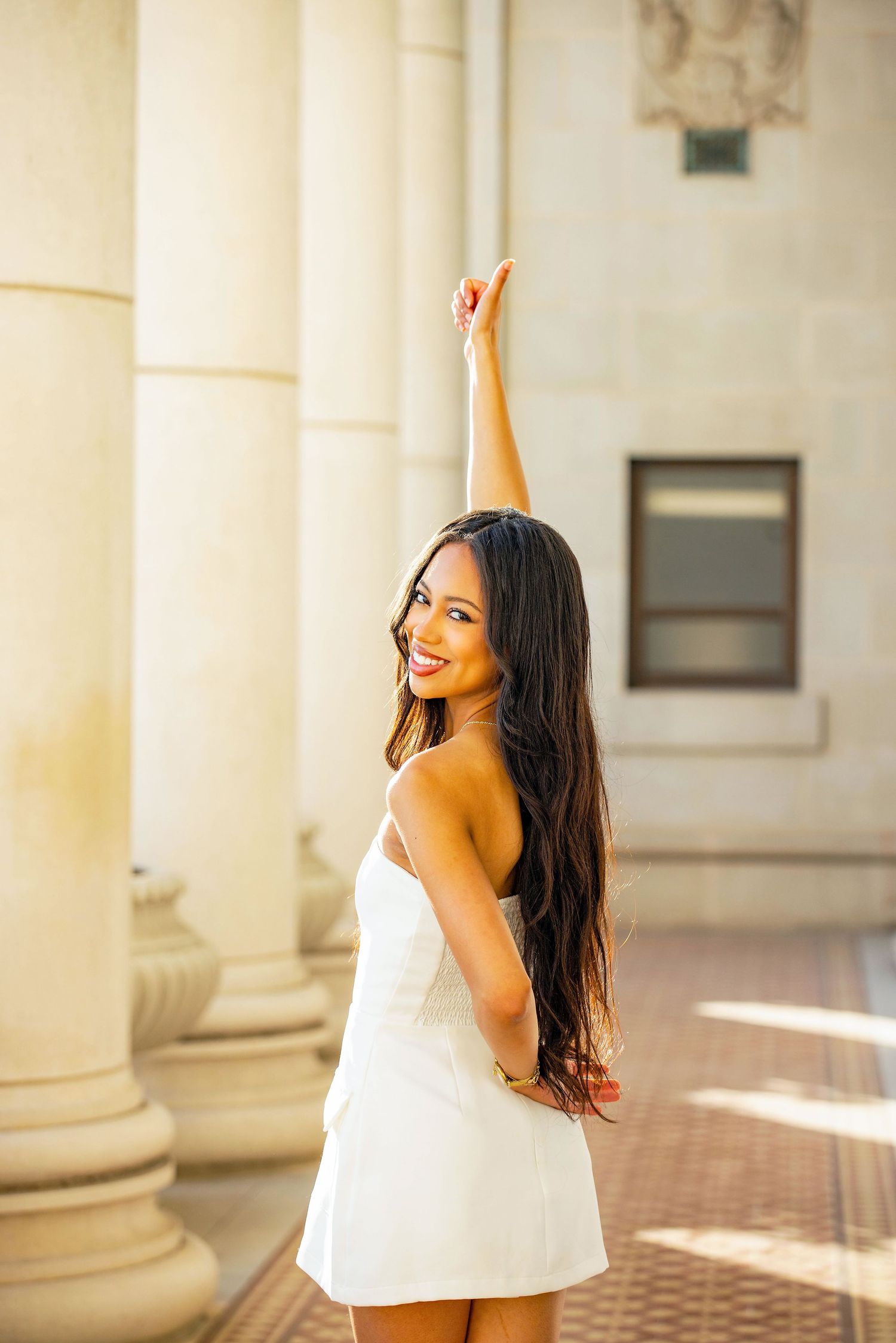 Woman in white dress smiling and posing against classical columns in warm sunlight.