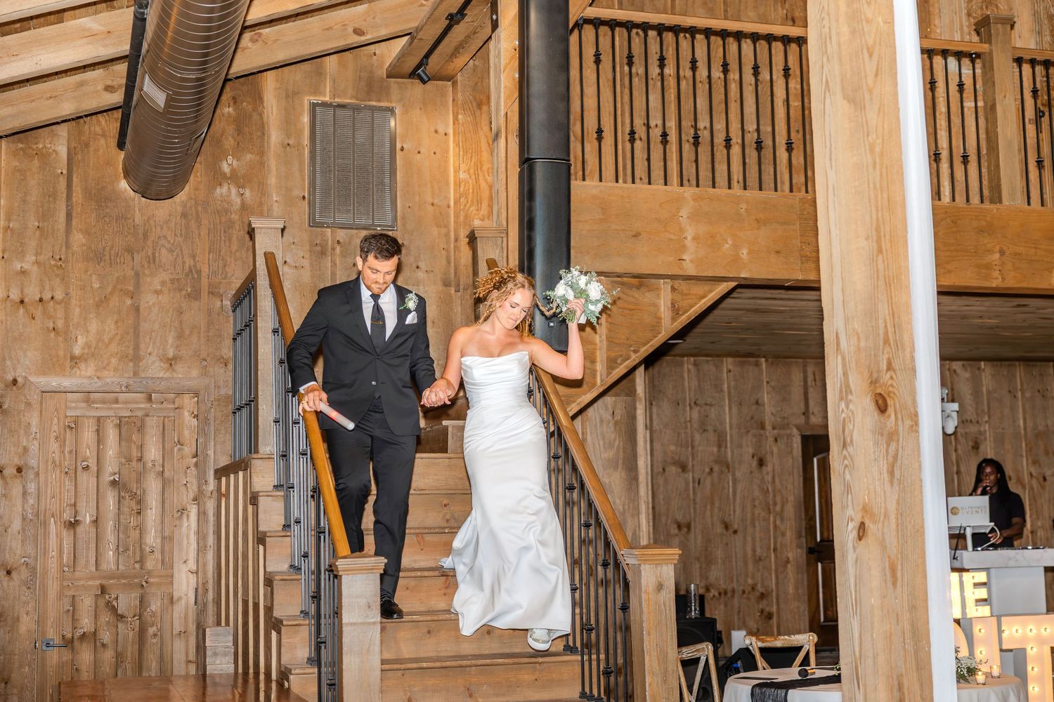 Bride and groom descend rustic wooden staircase in barn wedding venue, bride holding bouquet of white flowers.